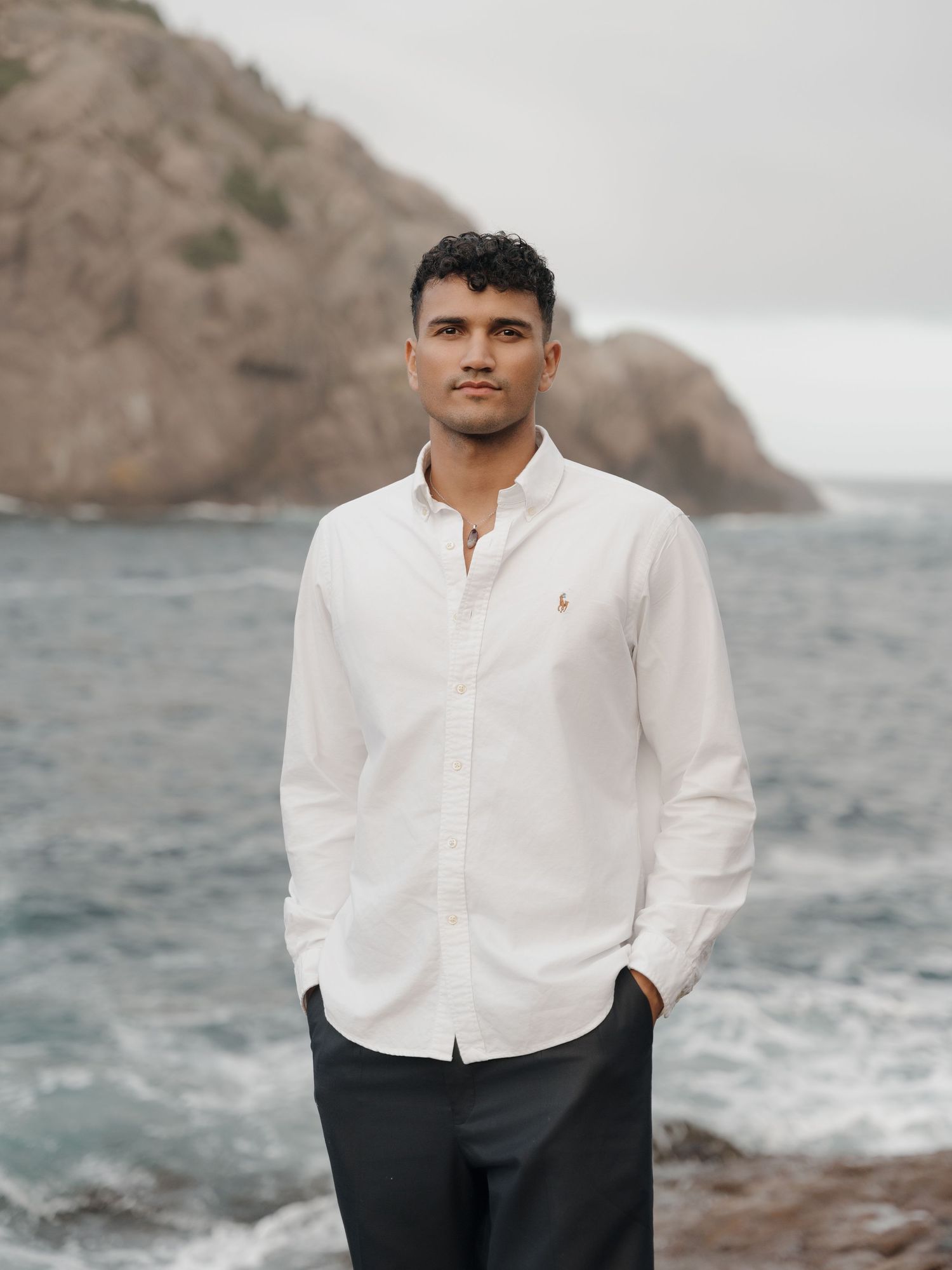 Person wearing white button-up shirt stands near rocky coastline with crashing waves in moody gray weather.