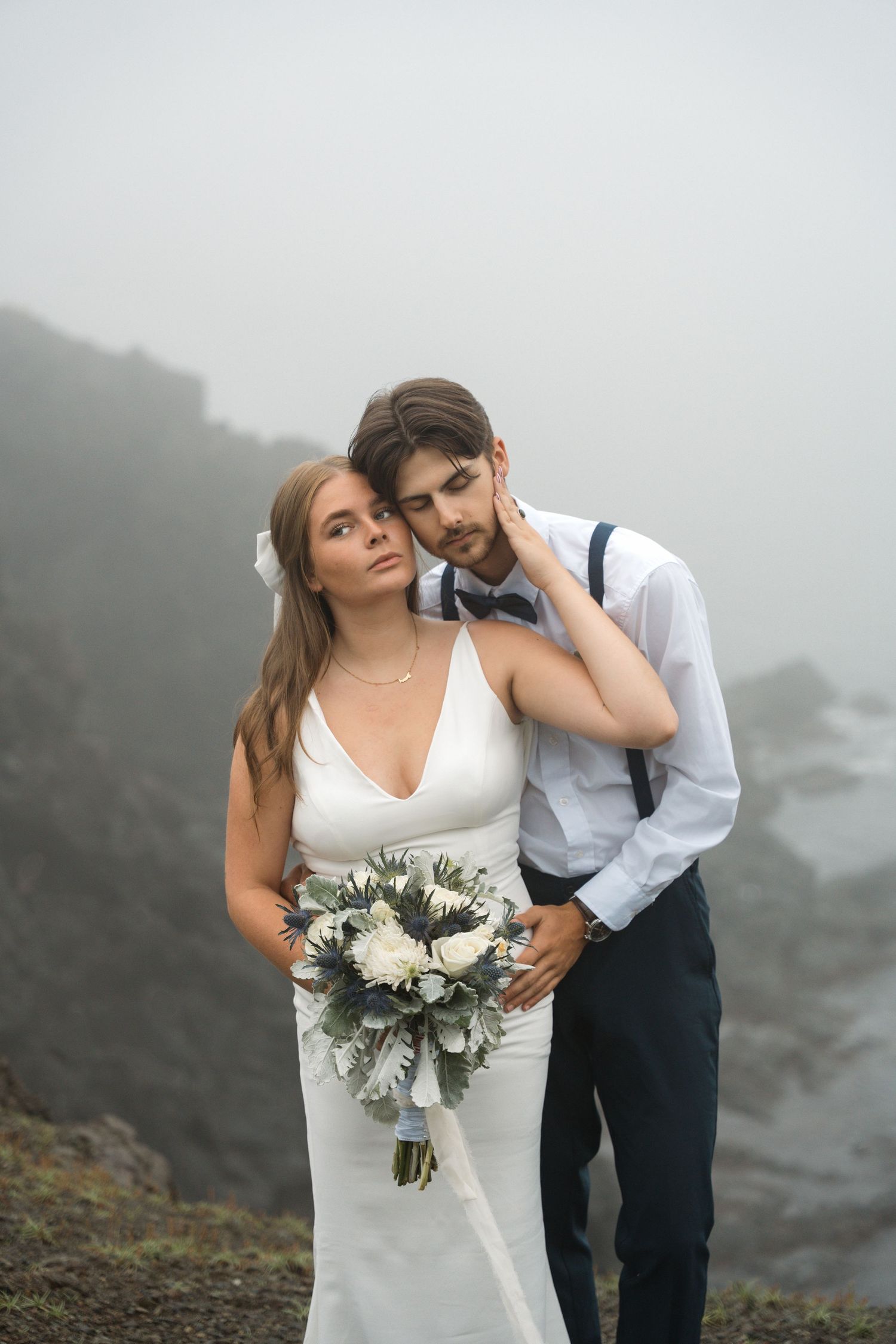 Wedding couple shares intimate moment on foggy mountainside while bride holds white and green floral bouquet.