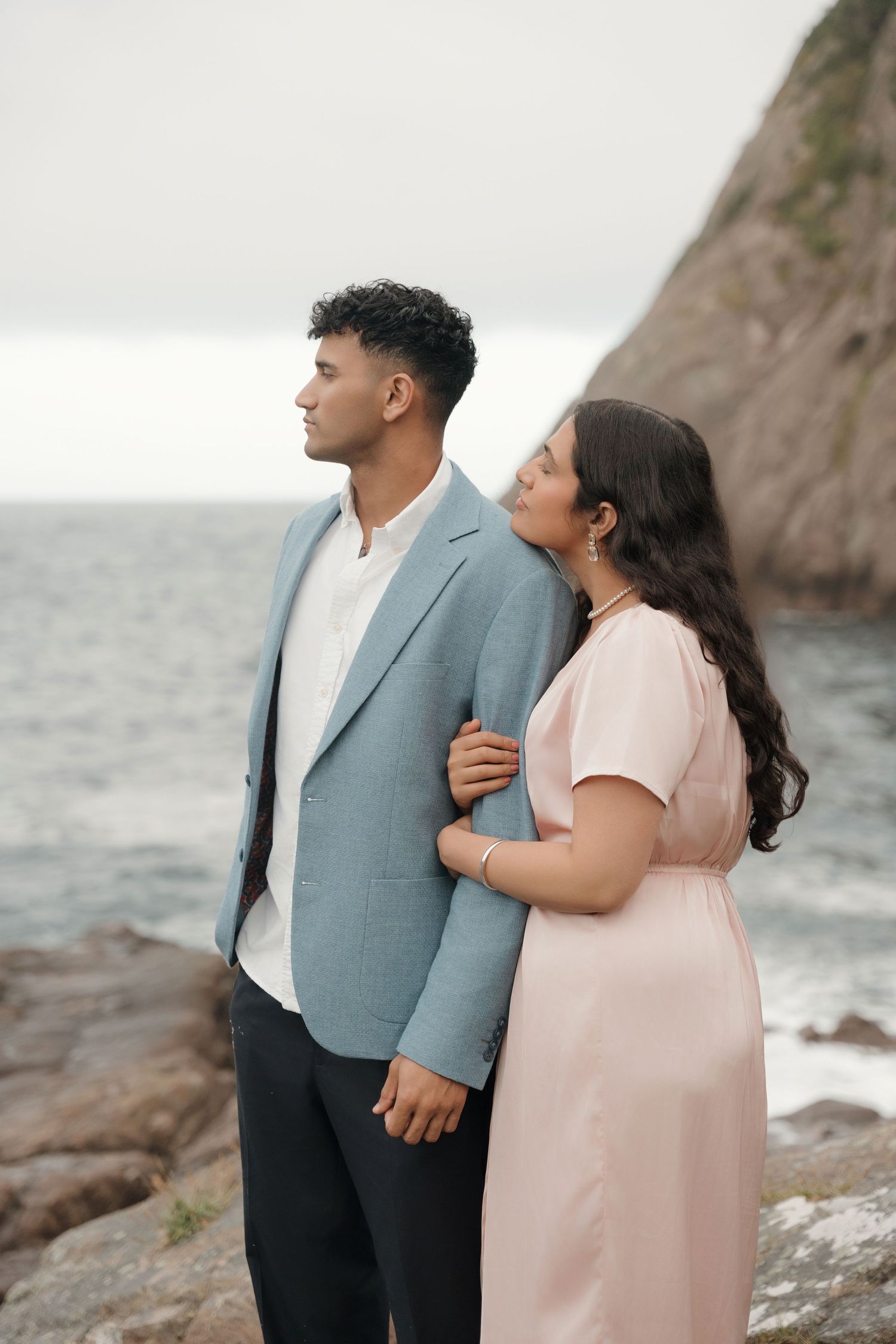 A couple in pink dress and gray suit embraces while standing on rocky coastal cliffs overlooking the ocean.