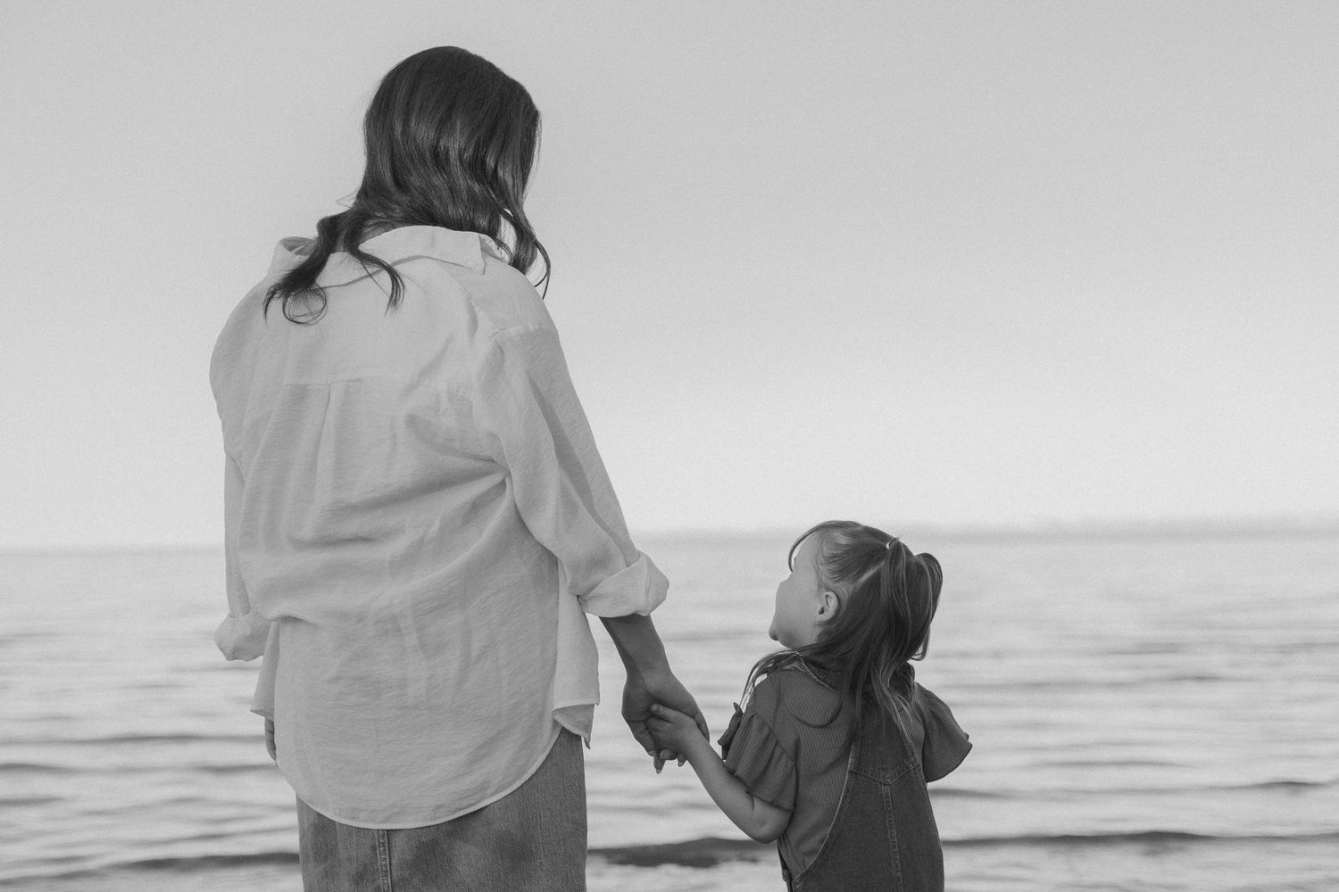 Parent and child hold hands while looking out at ocean during sunset.