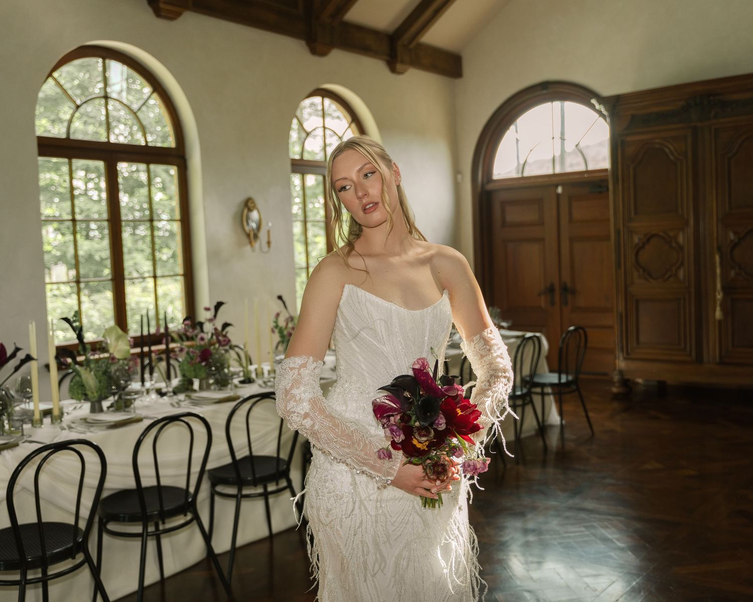Elegant figure in a lace wedding dress holds deep burgundy bouquet in rustic venue with arched windows and wooden chairs.