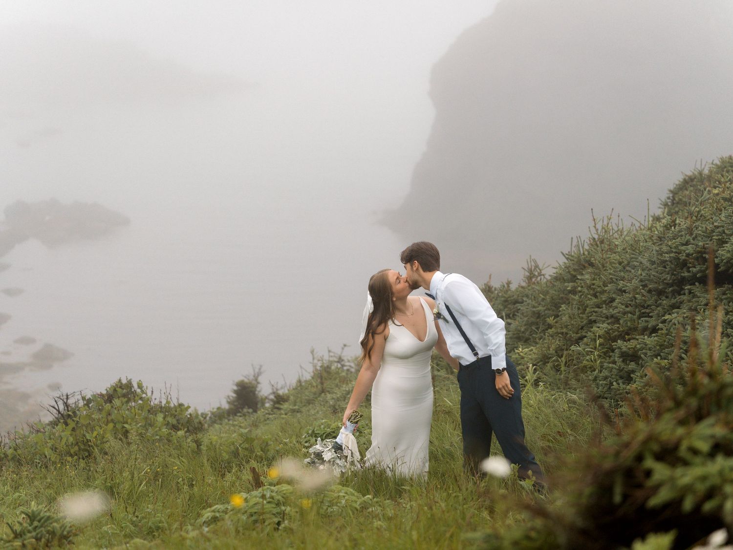 Wedding couple in formal attire stand together on a foggy mountain slope with greenery.