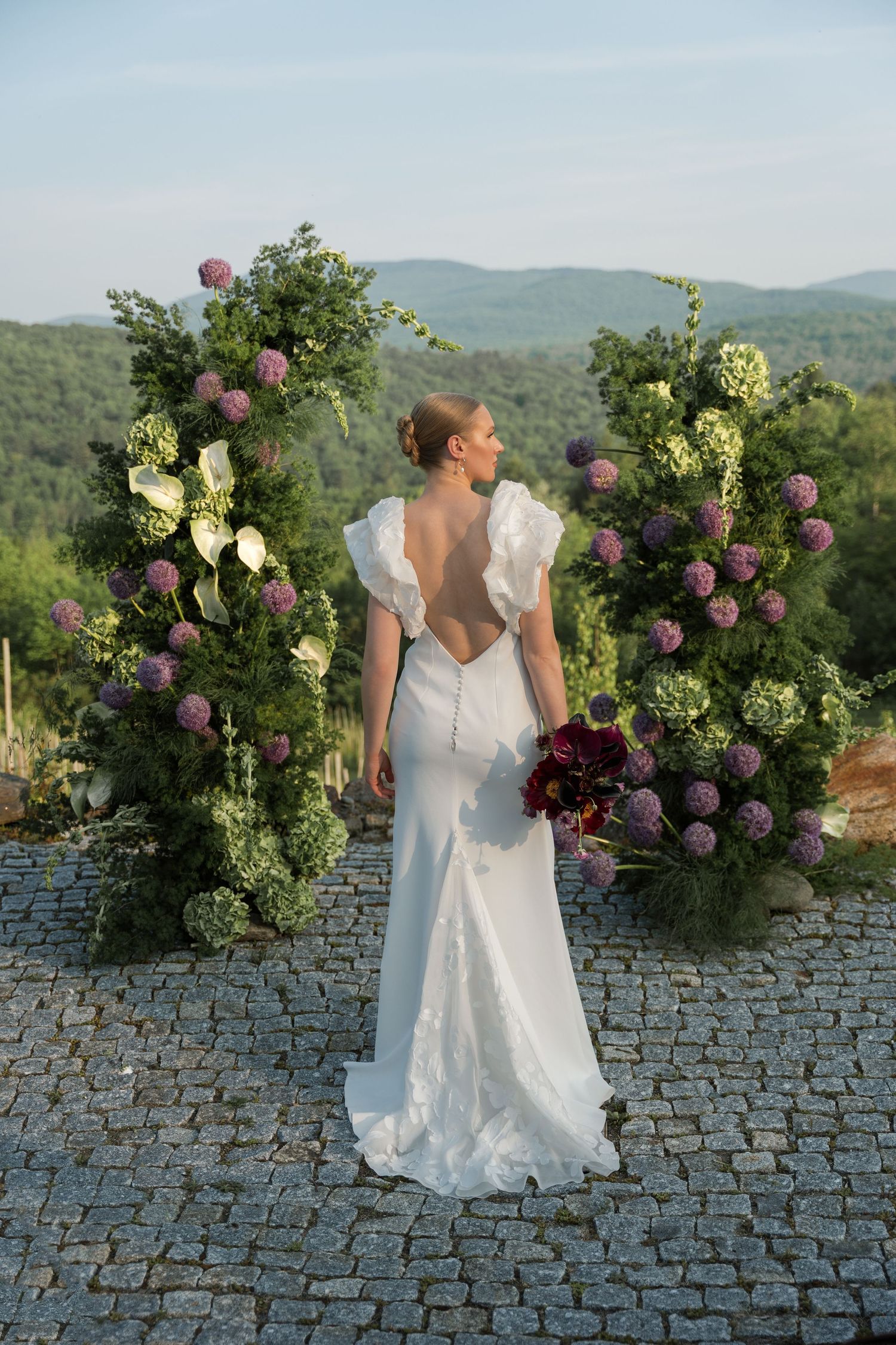 A wedding dress is captured against a stone wall adorned with purple and green florals in a mountain setting.