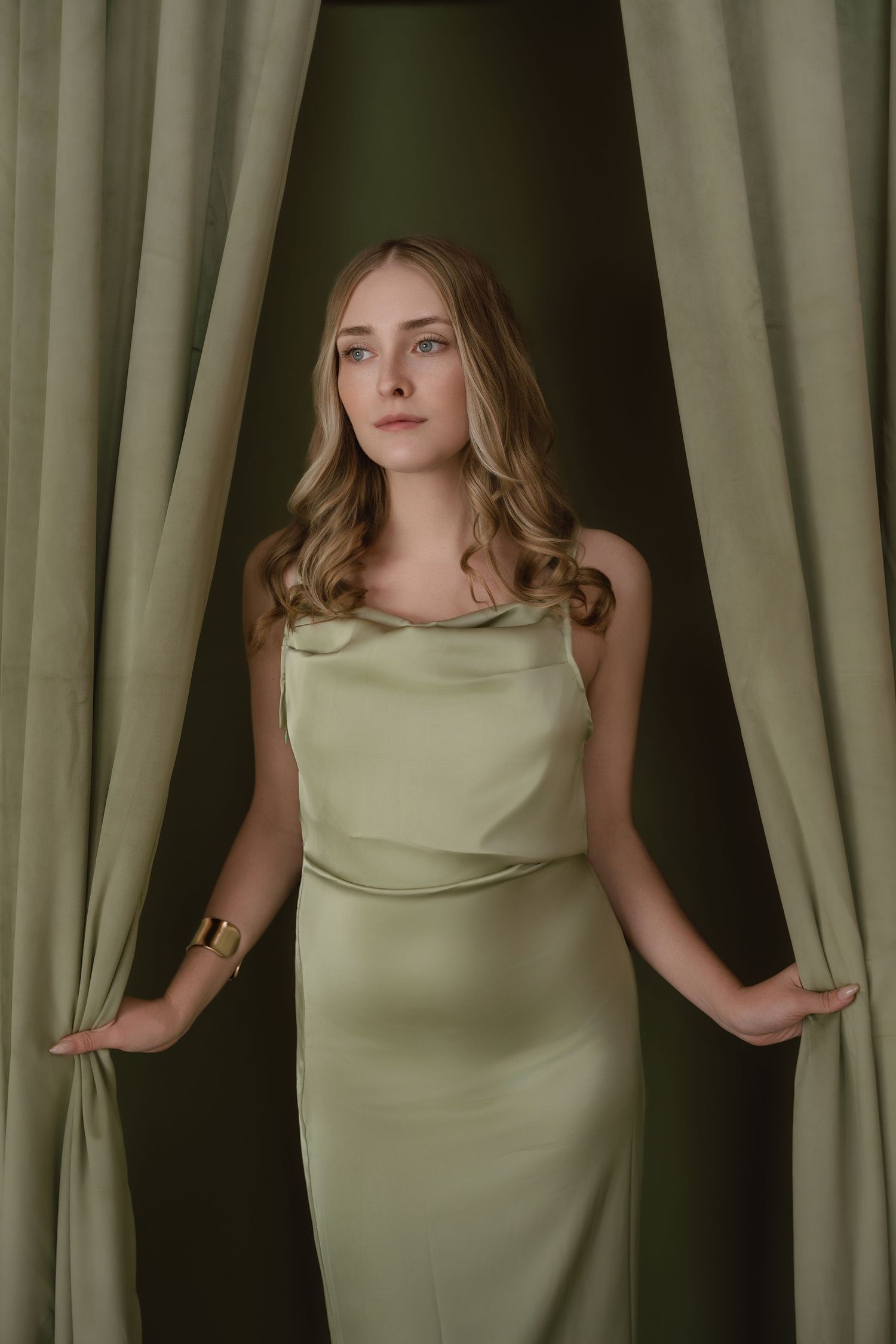 A woman poses in a light green sleeveless dress against a backdrop of olive curtains in a fashion photo series.