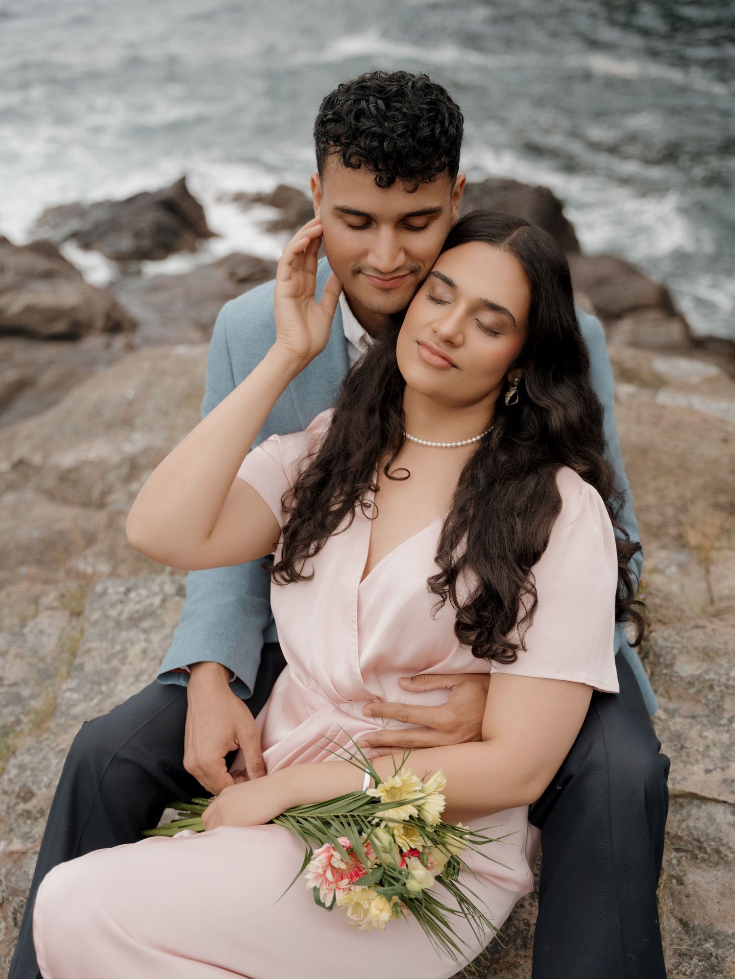 A romantic couple sits on coastal rocks near crashing waves as she holds a small bouquet in her pink dress.