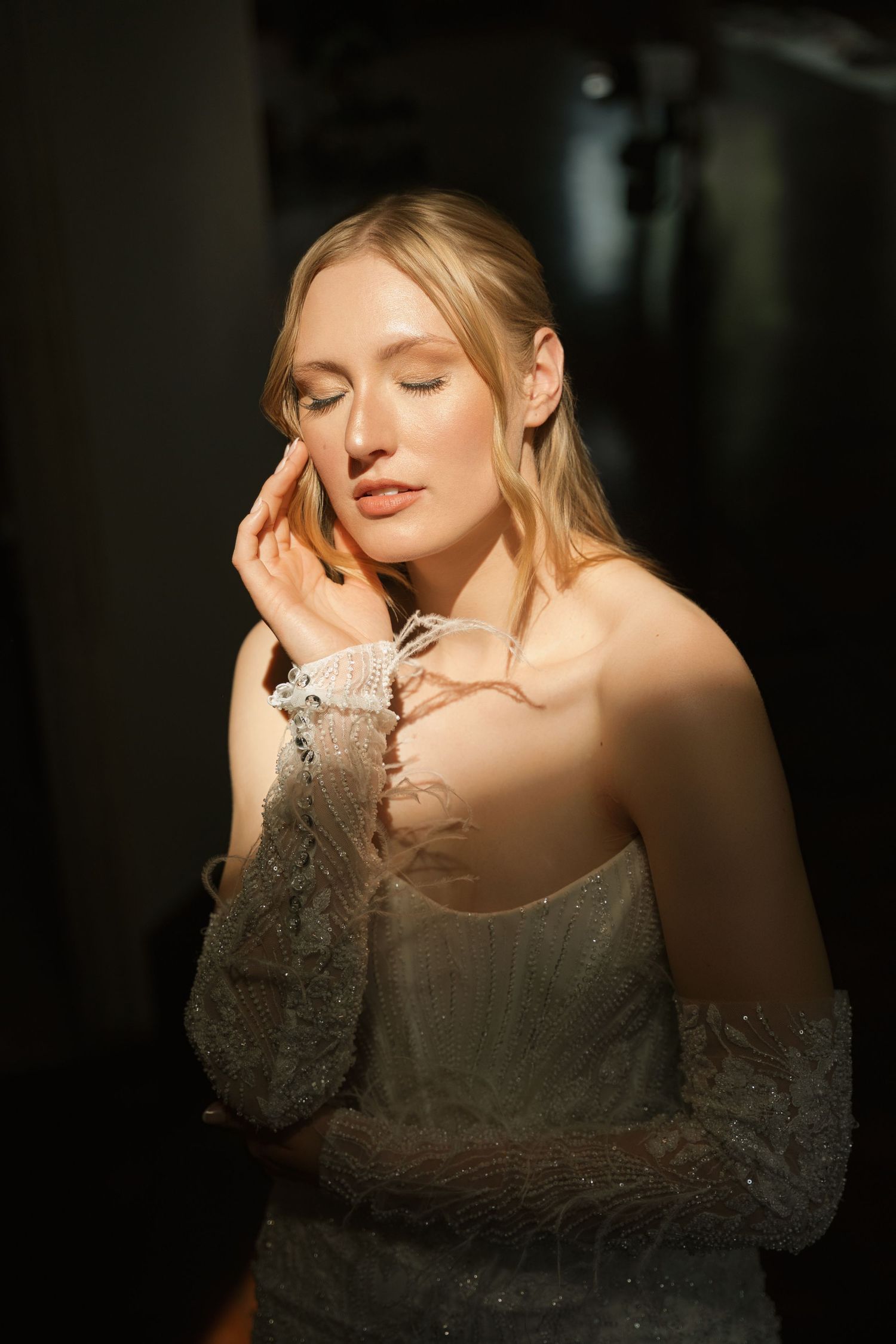 A person in a lace dress poses against a dark background with dramatic lighting and closed eyes.