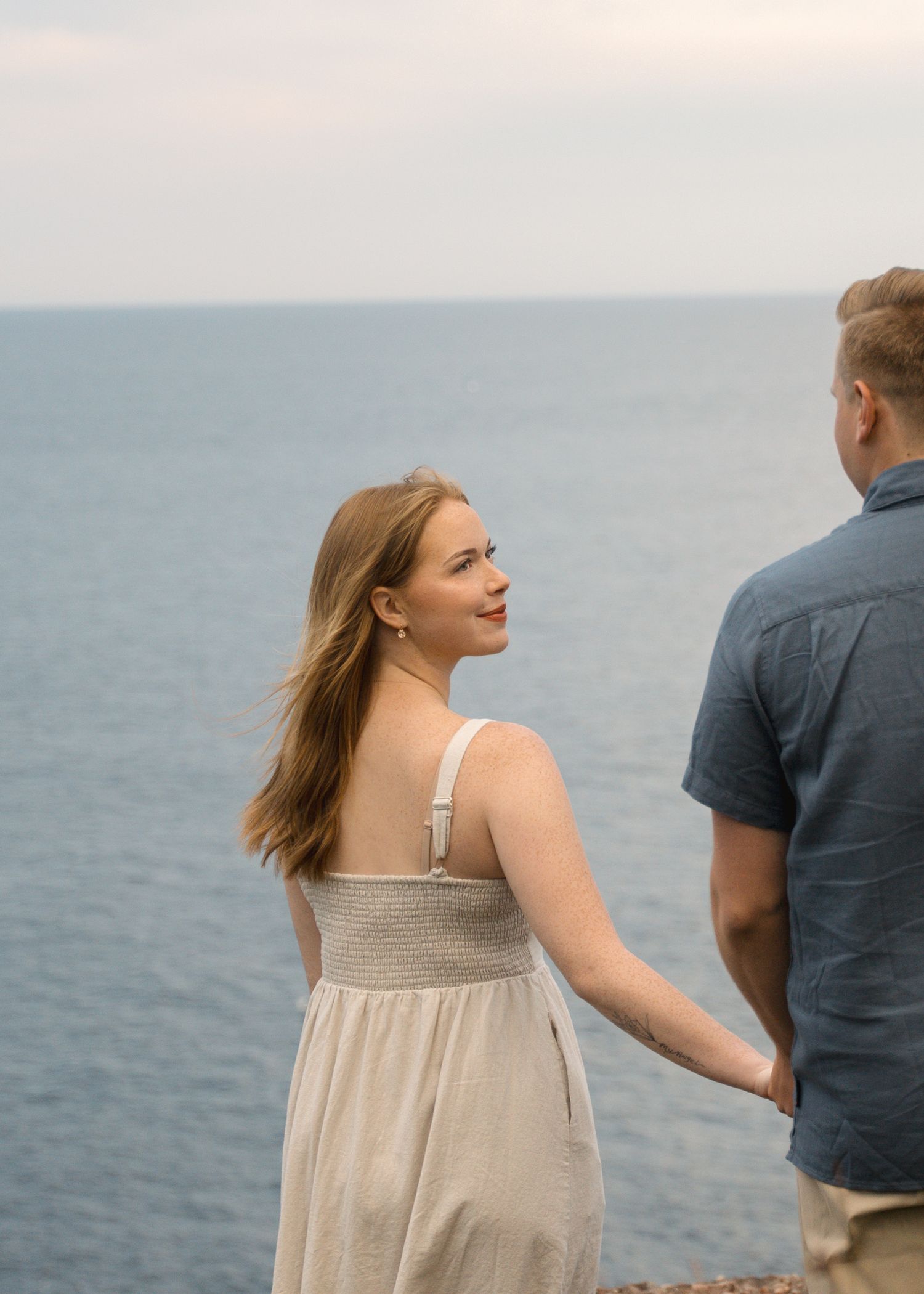 A couple stands together on a seaside cliff looking out at the ocean on a serene afternoon.