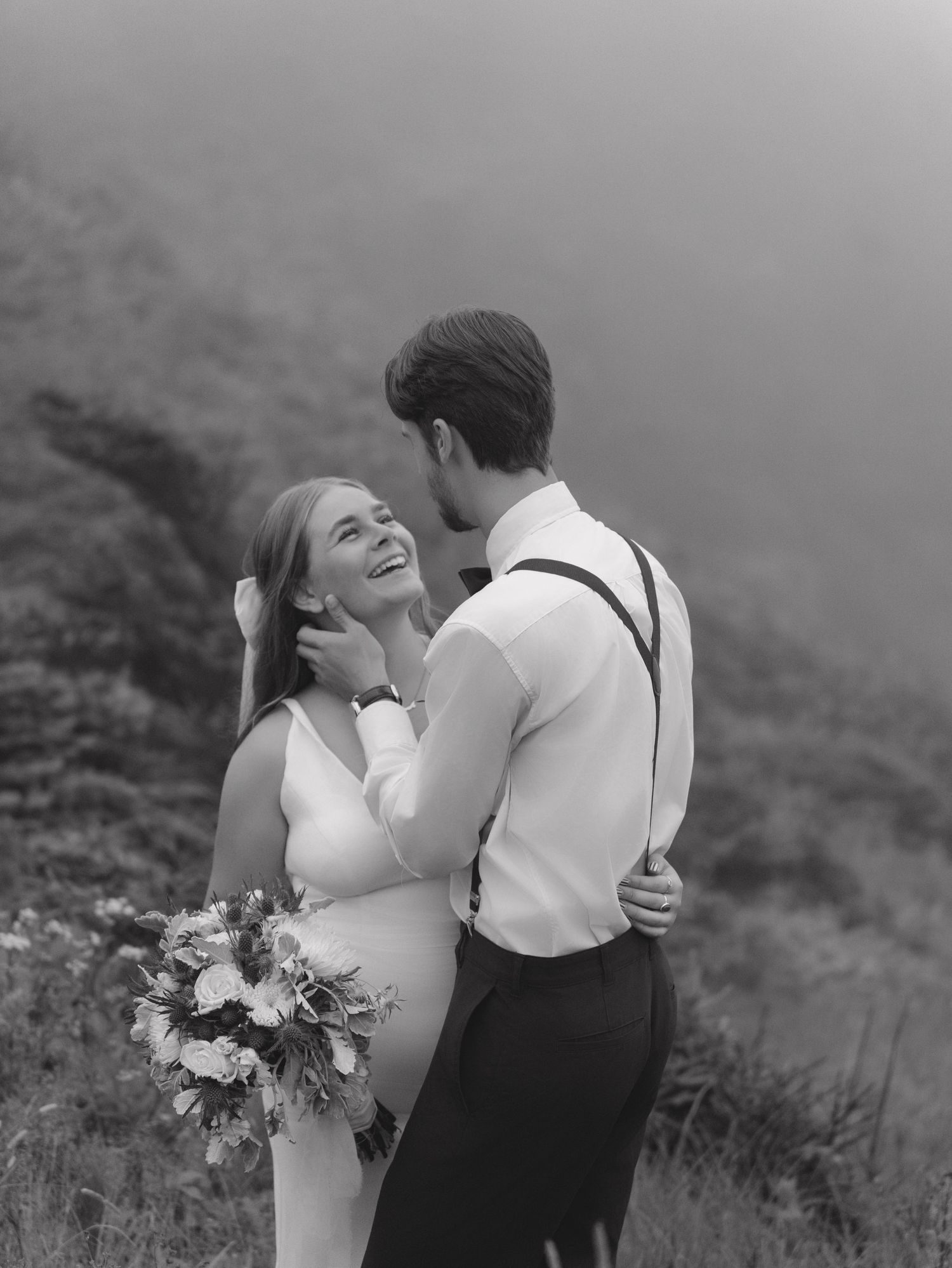 Wedding couple embraces and laughs together on a fog-covered mountainside in black and white.
