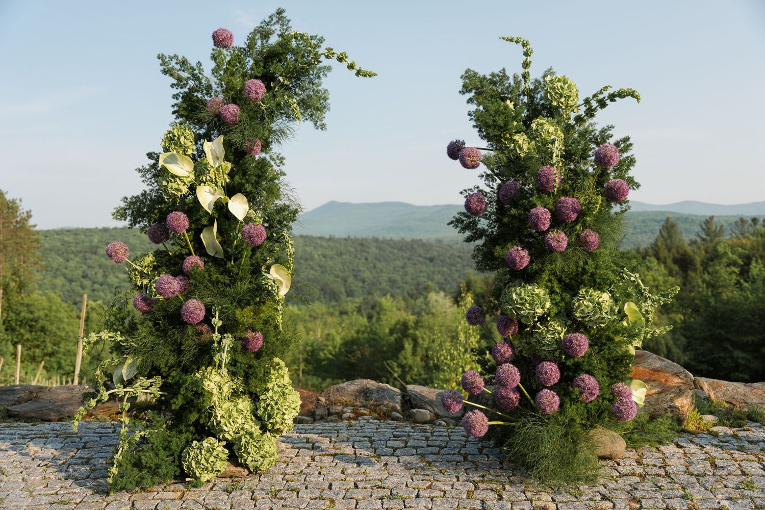 Rustic wine bottles decorated with pink and white flowers arranged along a stone wall with mountains in the backdrop.