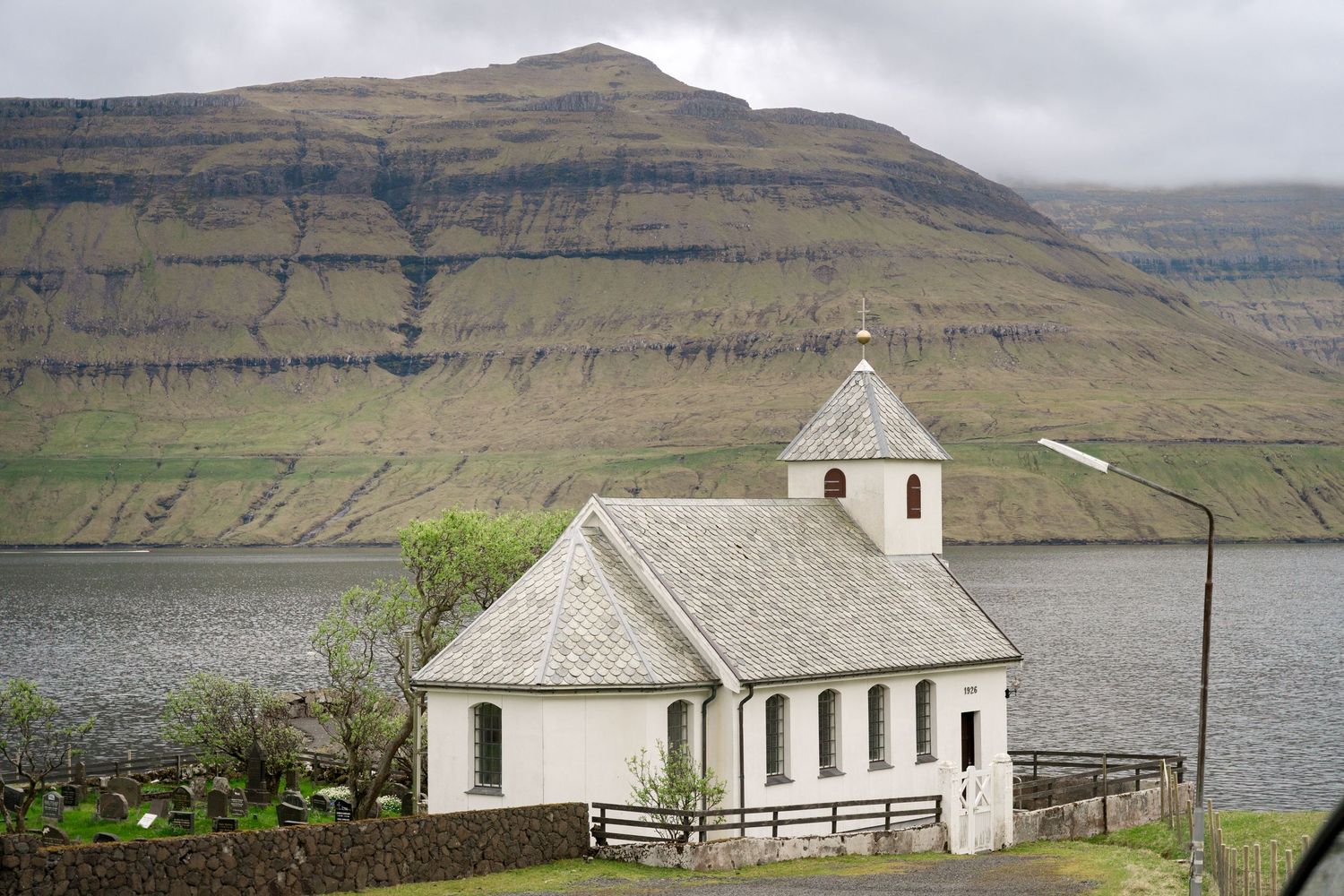 White church with bell tower beside a fjord, dramatic layered mountain backdrop, Faroe Islands landscape.