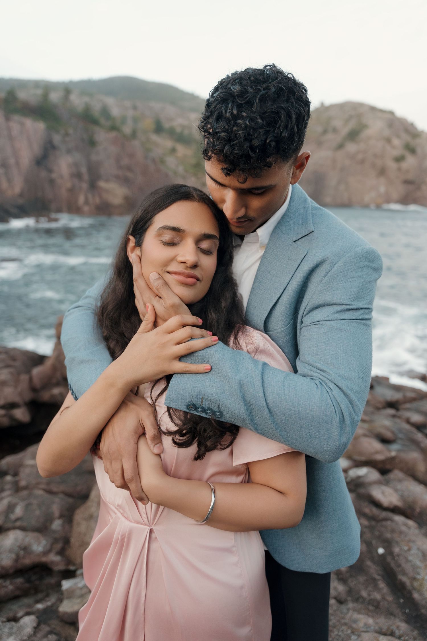 Romantic couple embraces on rocky coastal cliffs with ocean waves in the background creating intimate moments.