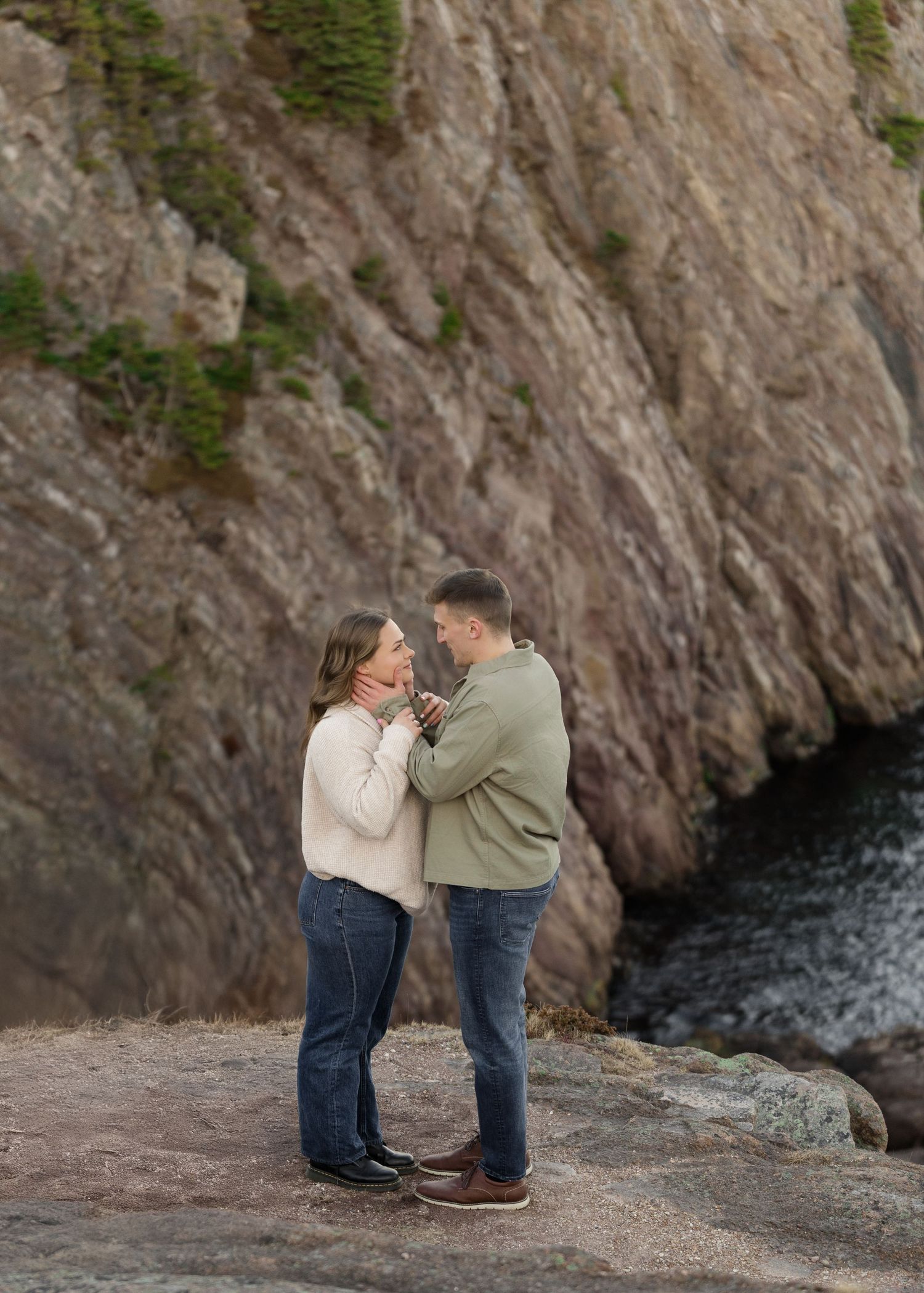 A couple shares an intimate moment on a rocky cliff overlooking the ocean with a dramatic stone wall backdrop.