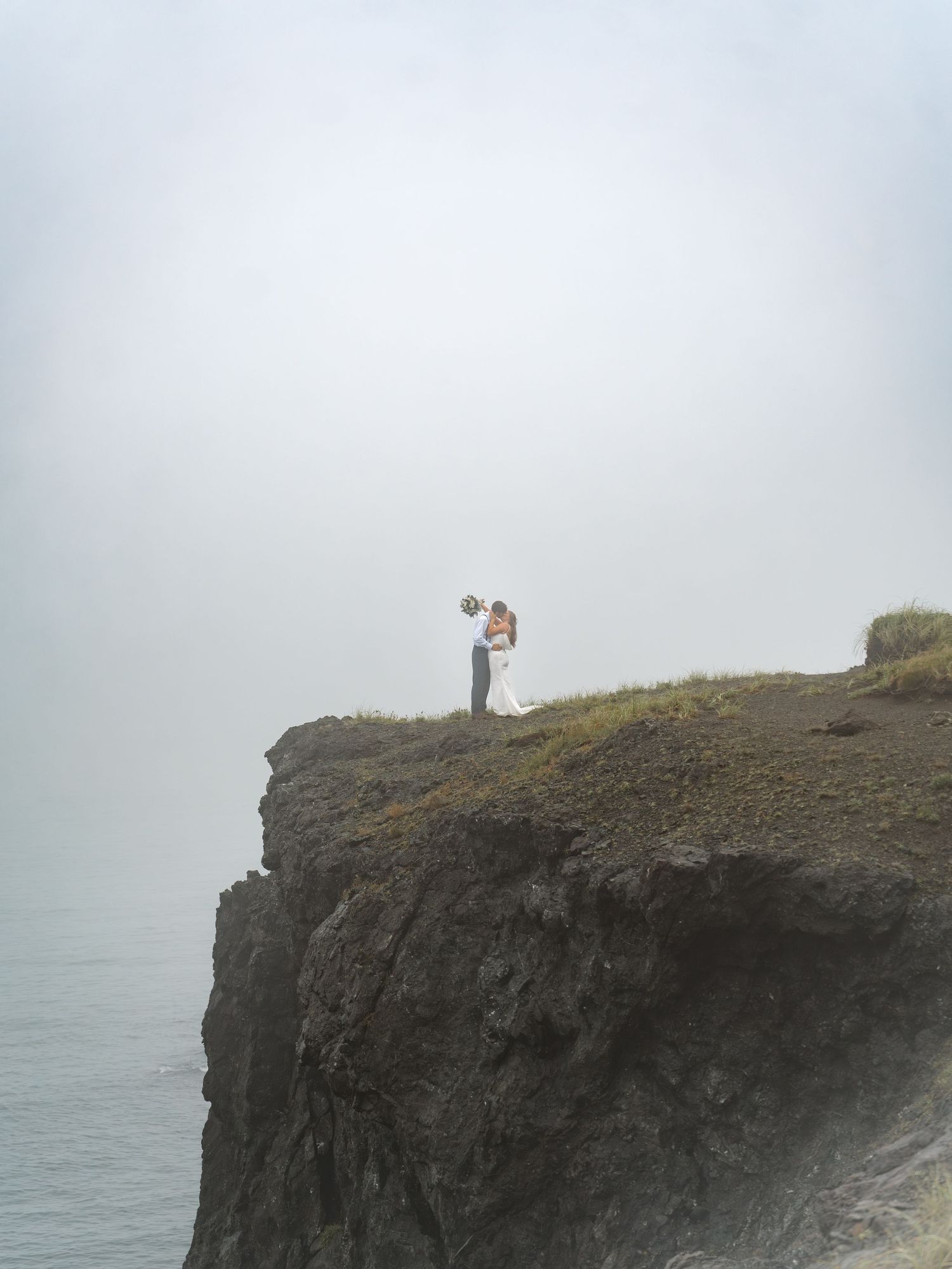 Dramatic coastal cliffs shrouded in fog overlooking the ocean with a small lighthouse in the distance.