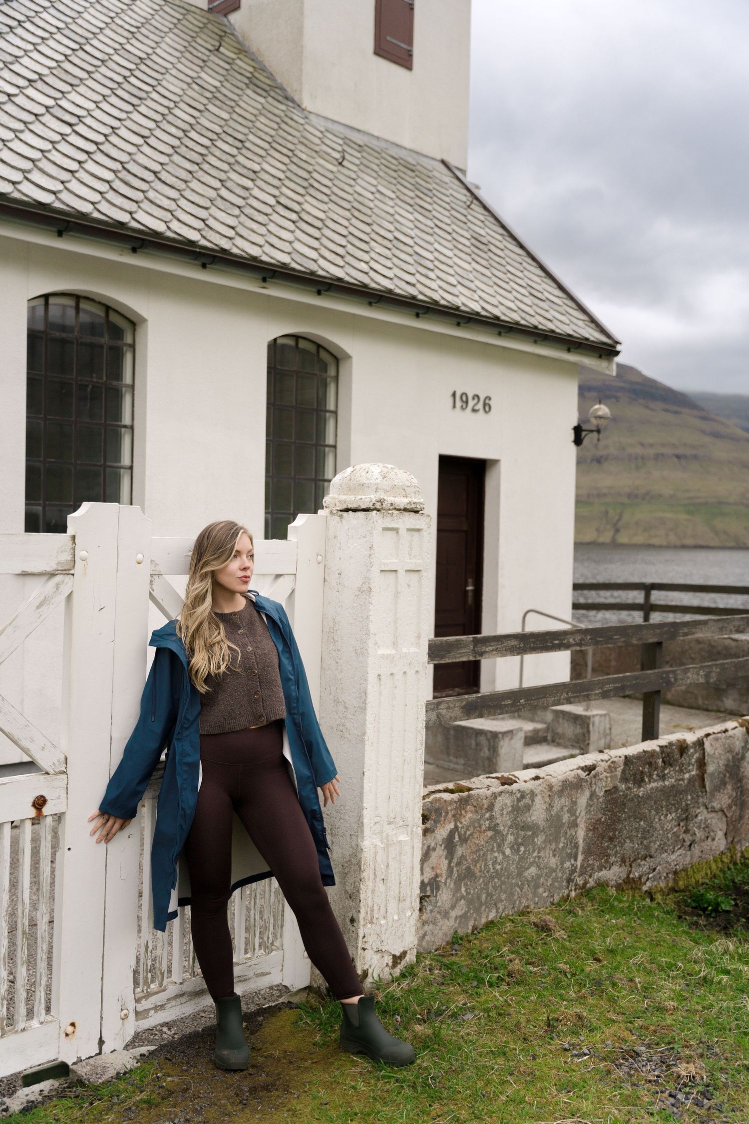 Woman in blue raincoat and burgundy leggings leaning against white gate of historic 1926 church in Iceland.
