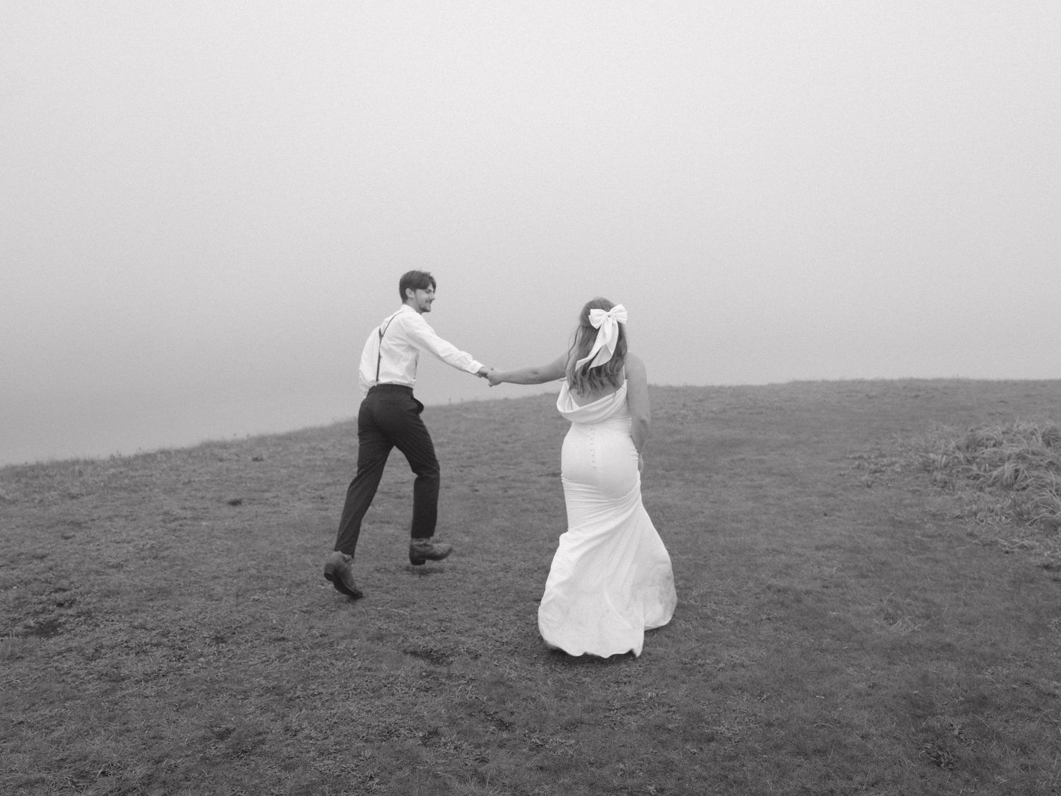 Newlyweds dance playfully together on a misty mountain ridge in black and white.