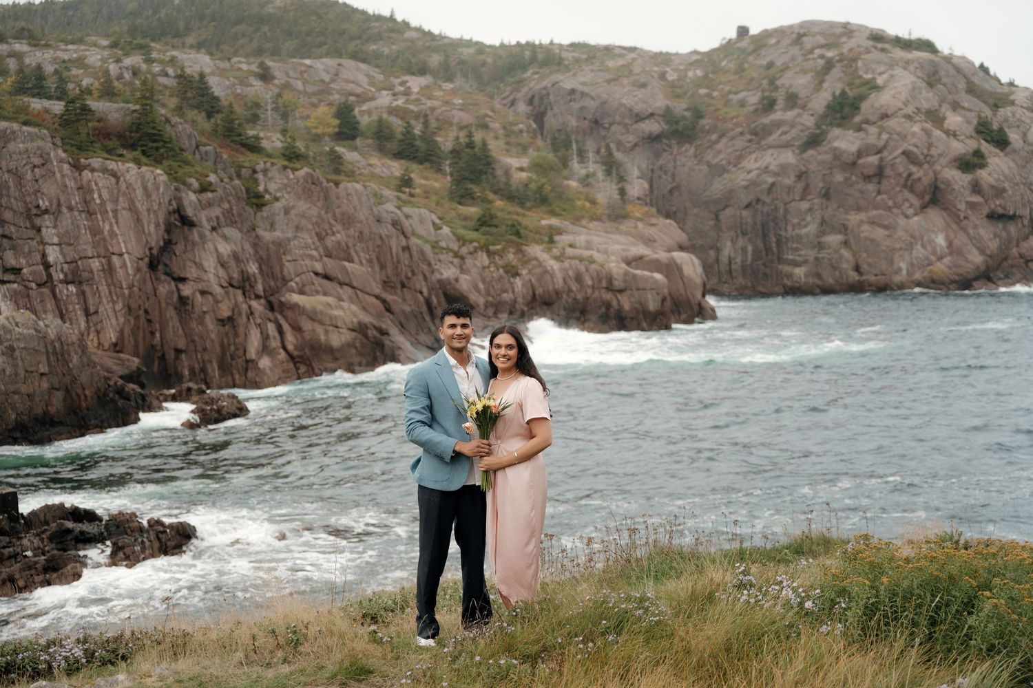 Couple standing on rocky coastal cliffs overlooking rugged ocean shoreline and crashing waves during golden hour.