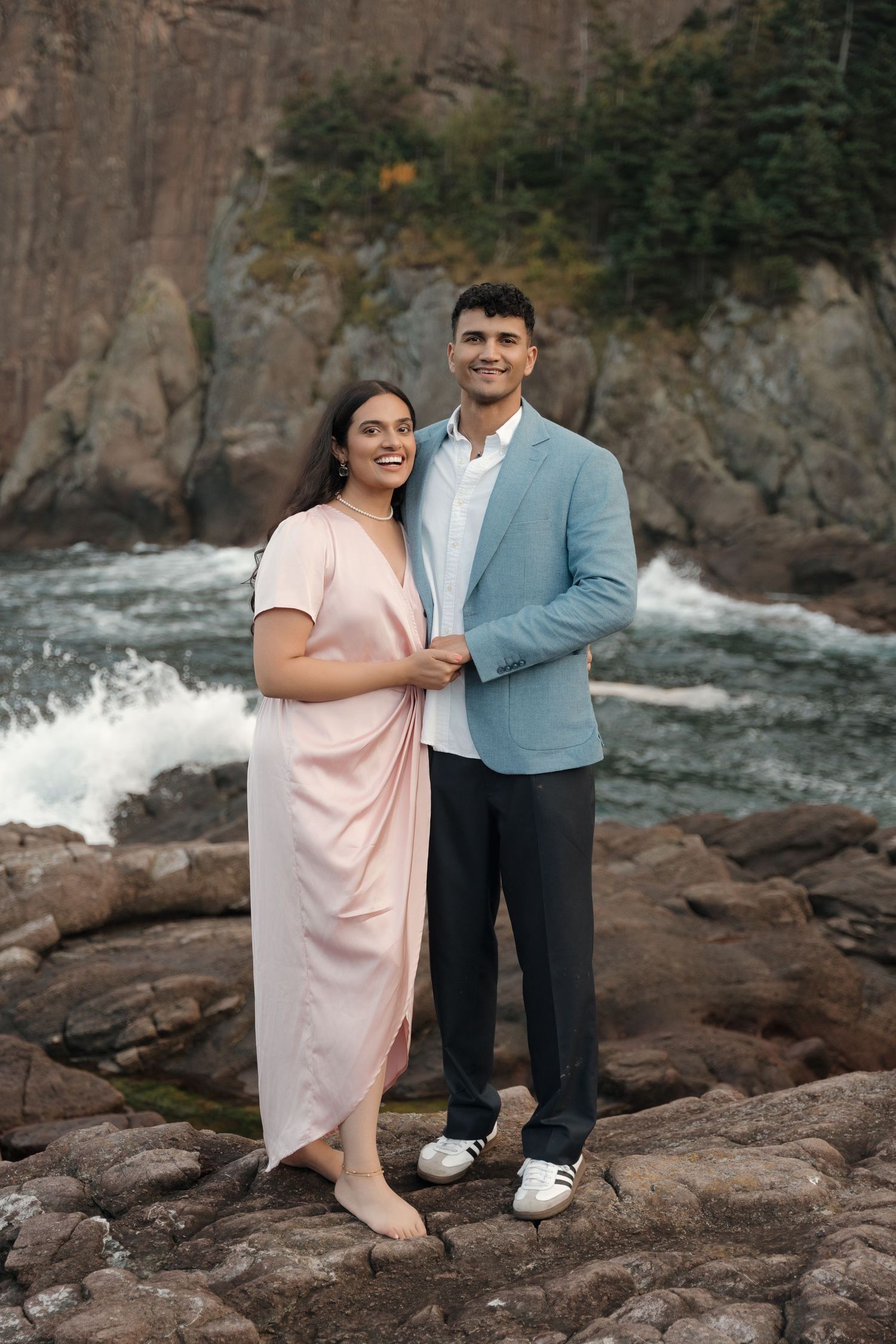 A couple poses together on rocky coastal cliffs beside a rushing river in an outdoor photoshoot.