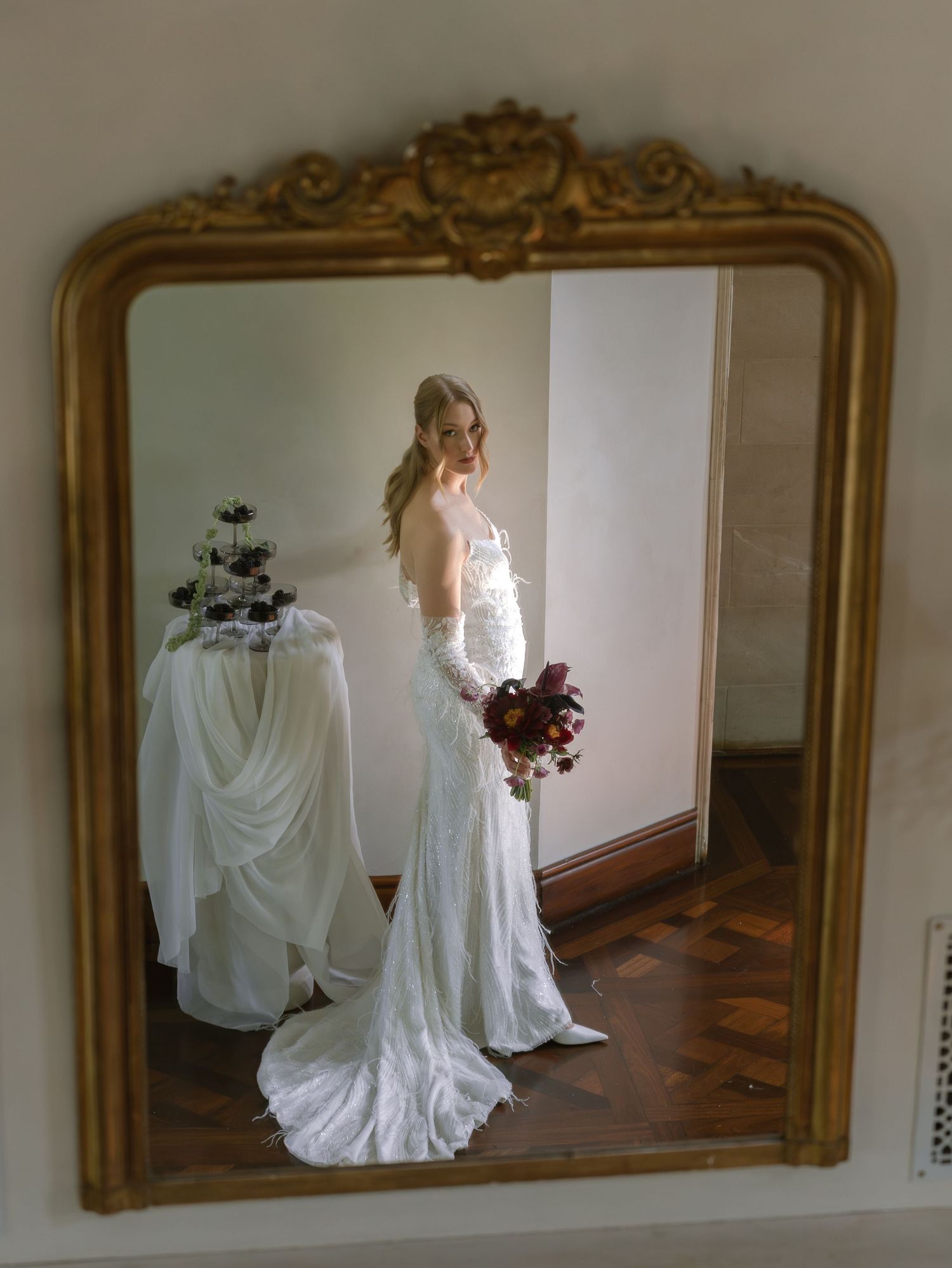 A figure in a flowing white vintage-style dress stands before an ornate gold-framed mirror in a sunlit room.