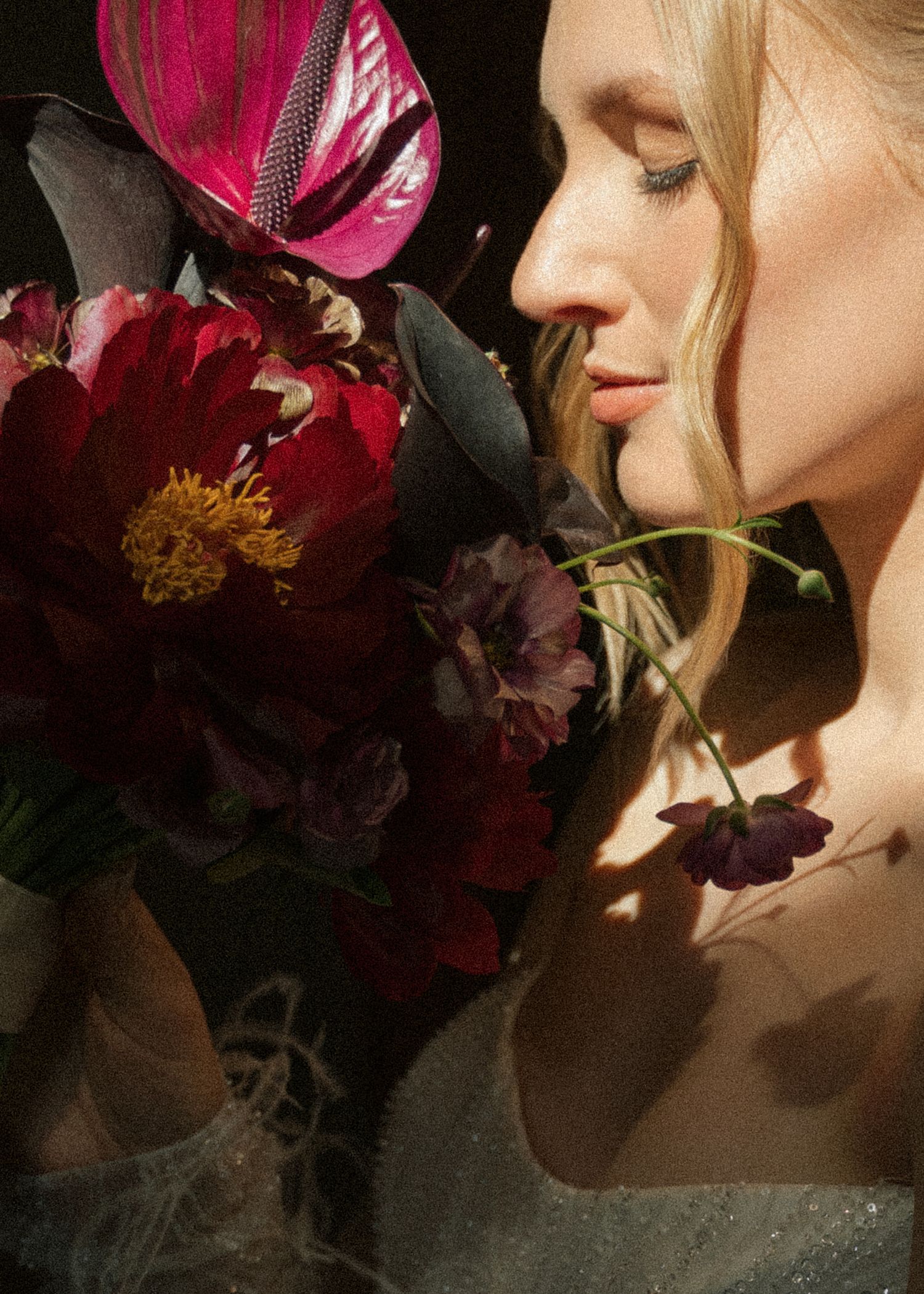 A woman with a pink fascinator poses artistically in front of deep red flowers creating a dramatic profile silhouette.