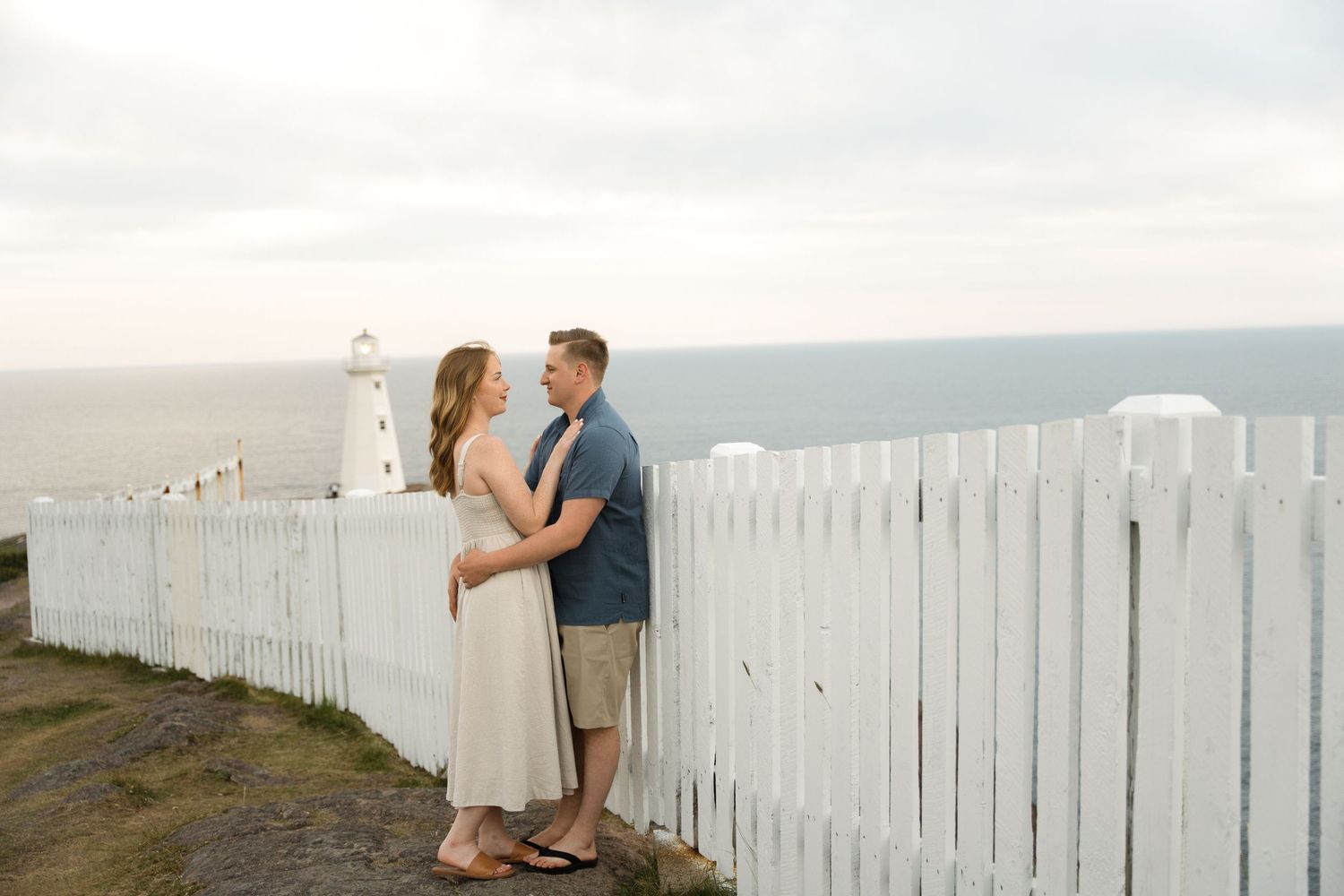 A romantic couple embraces near a white picket fence overlooking a coastal landscape at sunset.