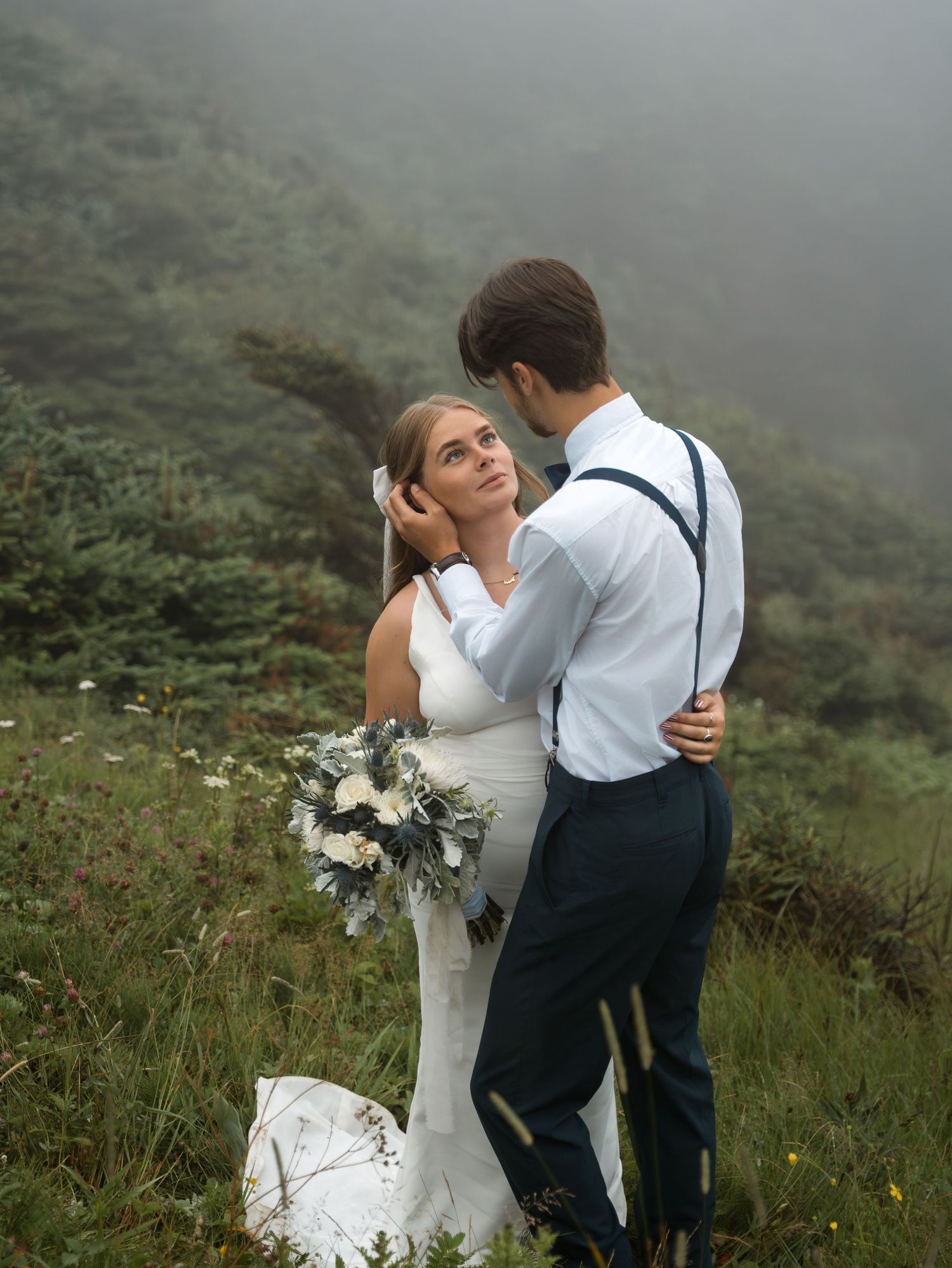 A couple shares an intimate moment in formal wear against a misty mountainous landscape with white flowers.