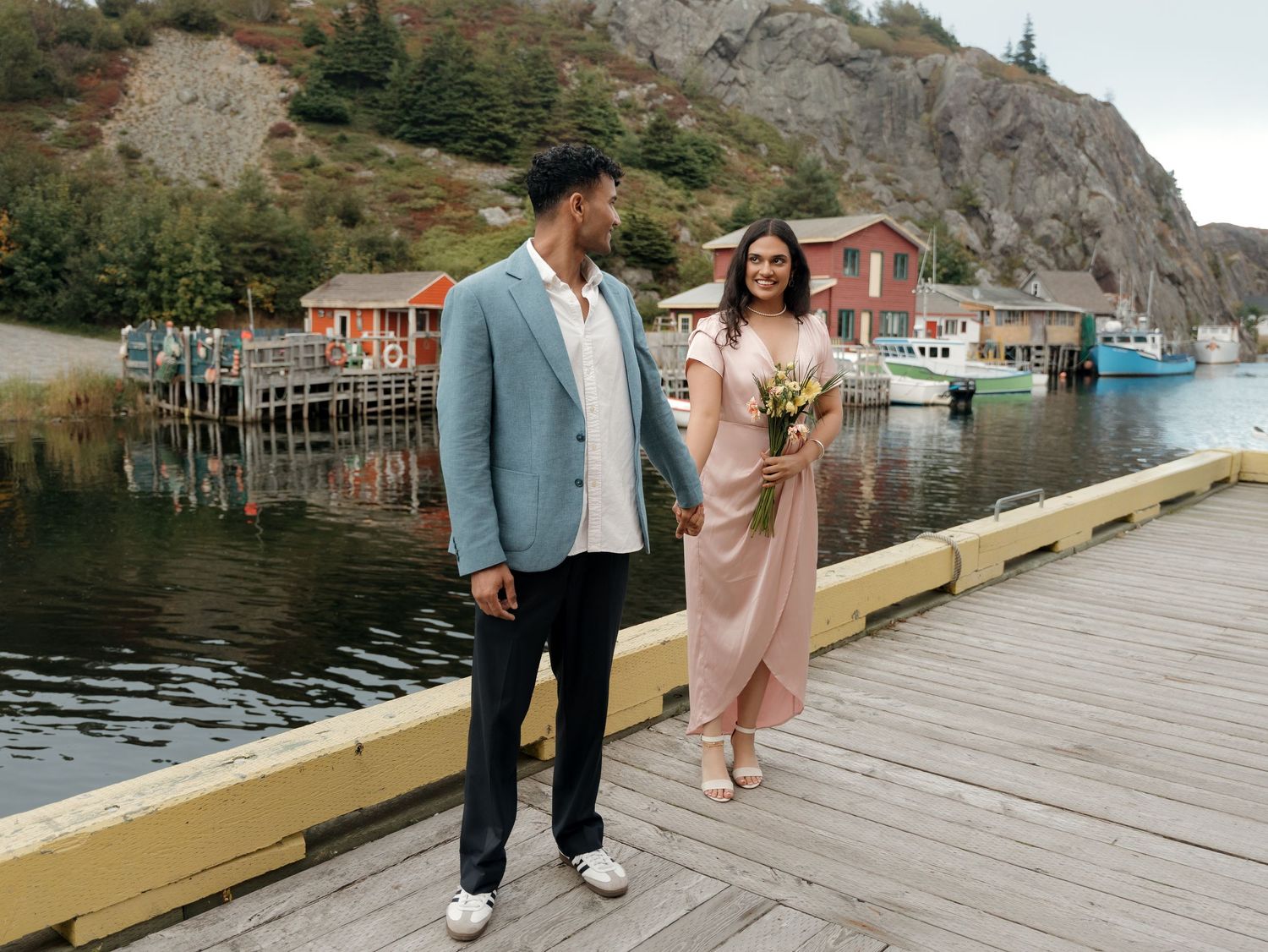 Person wearing a suit jacket walks beside someone in a pink dress on a wooden dock by a waterfront mountain view.