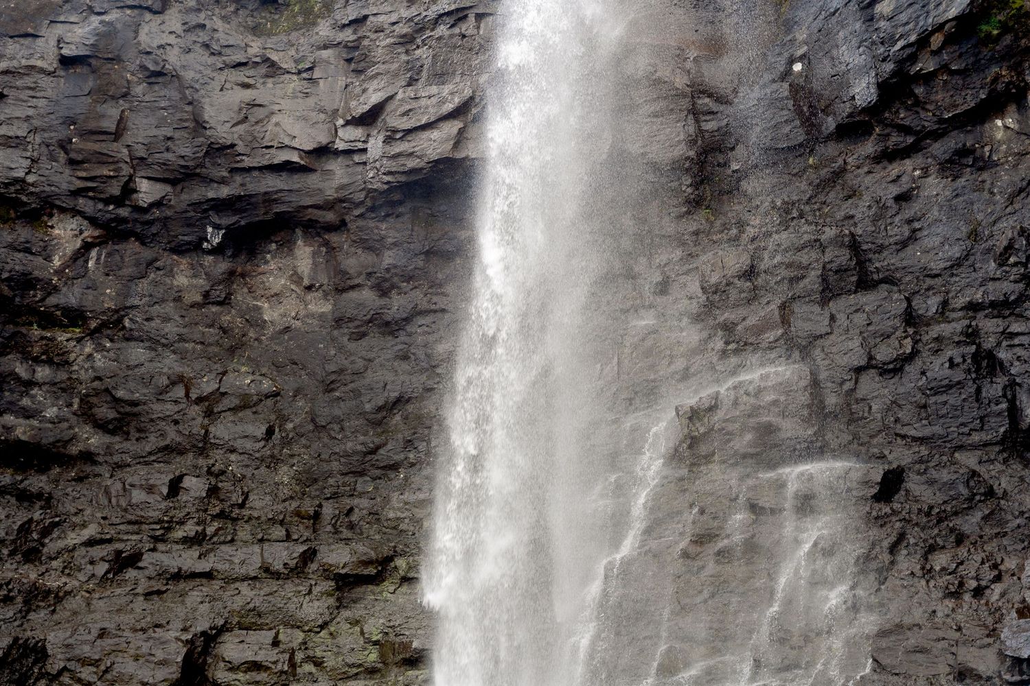A powerful waterfall cascades down a dark rocky cliff face, with mist rising from the rushing white water.