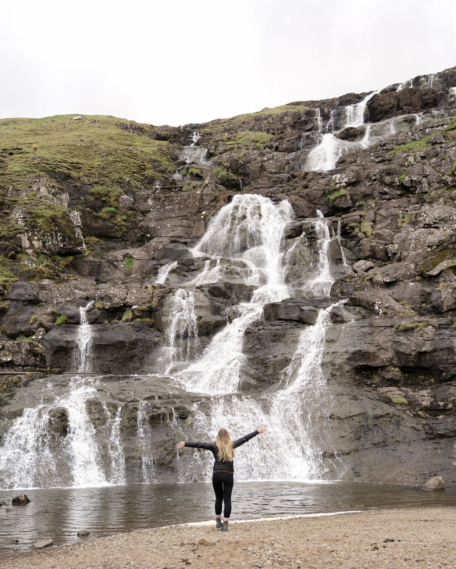 Woman standing arms outstretched before a stunning multi-tiered waterfall cascading down rocky cliffs in Iceland.