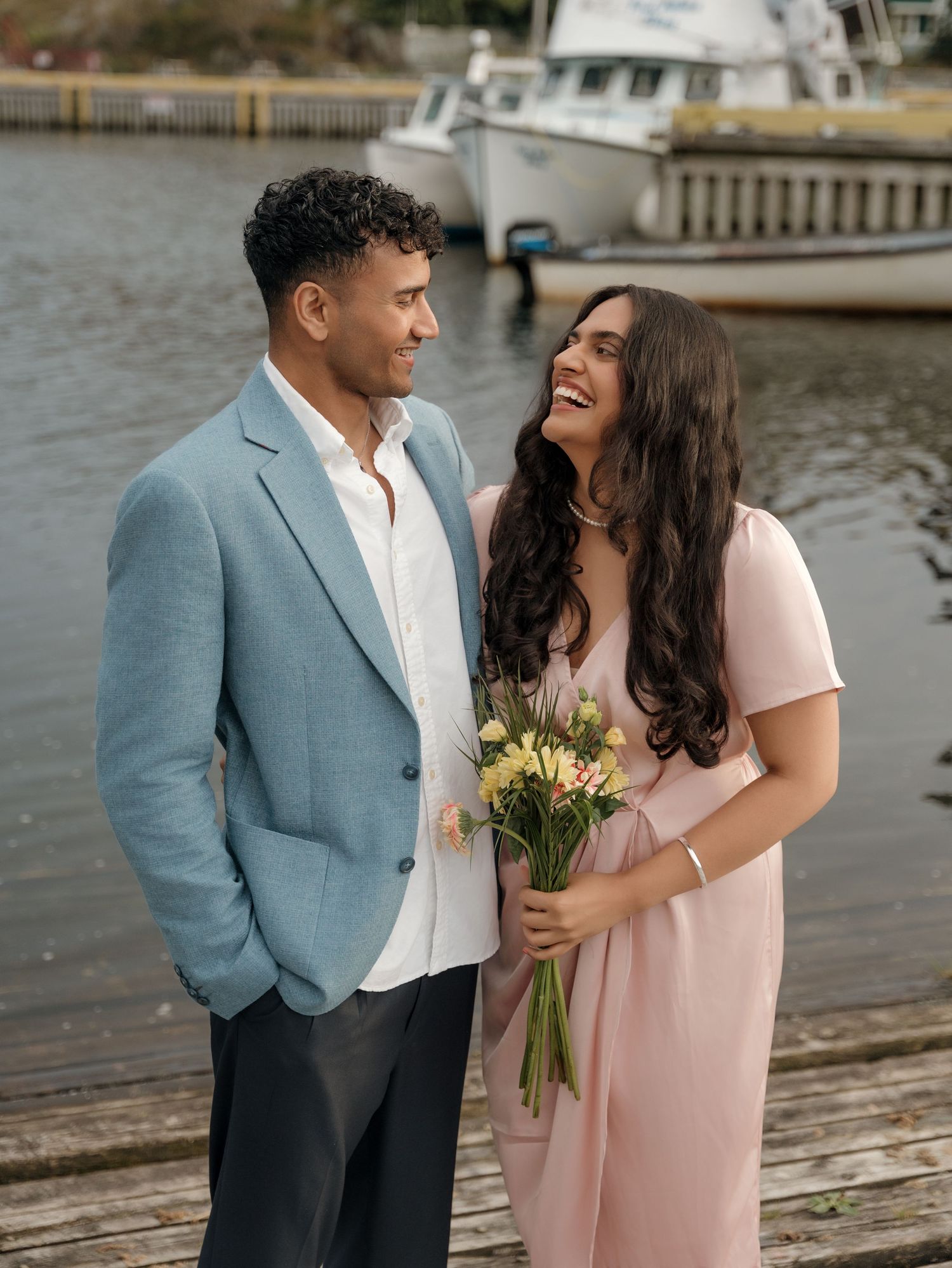 A couple shares a joyful moment on a dock wearing pink and blue formal attire while holding yellow flowers.