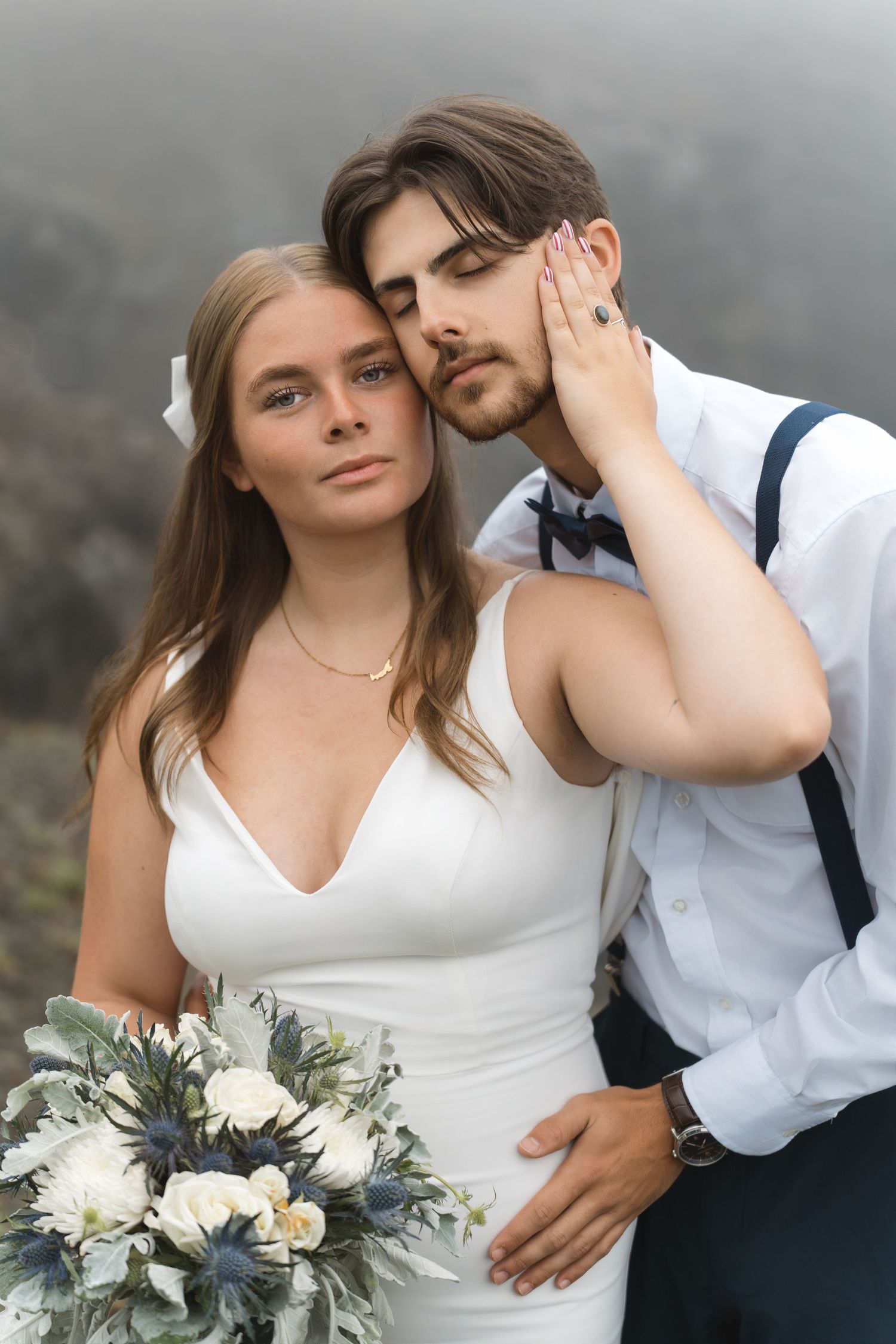 Romantic wedding couple poses together on misty mountain cliffs with elegant white and blue floral bouquet.