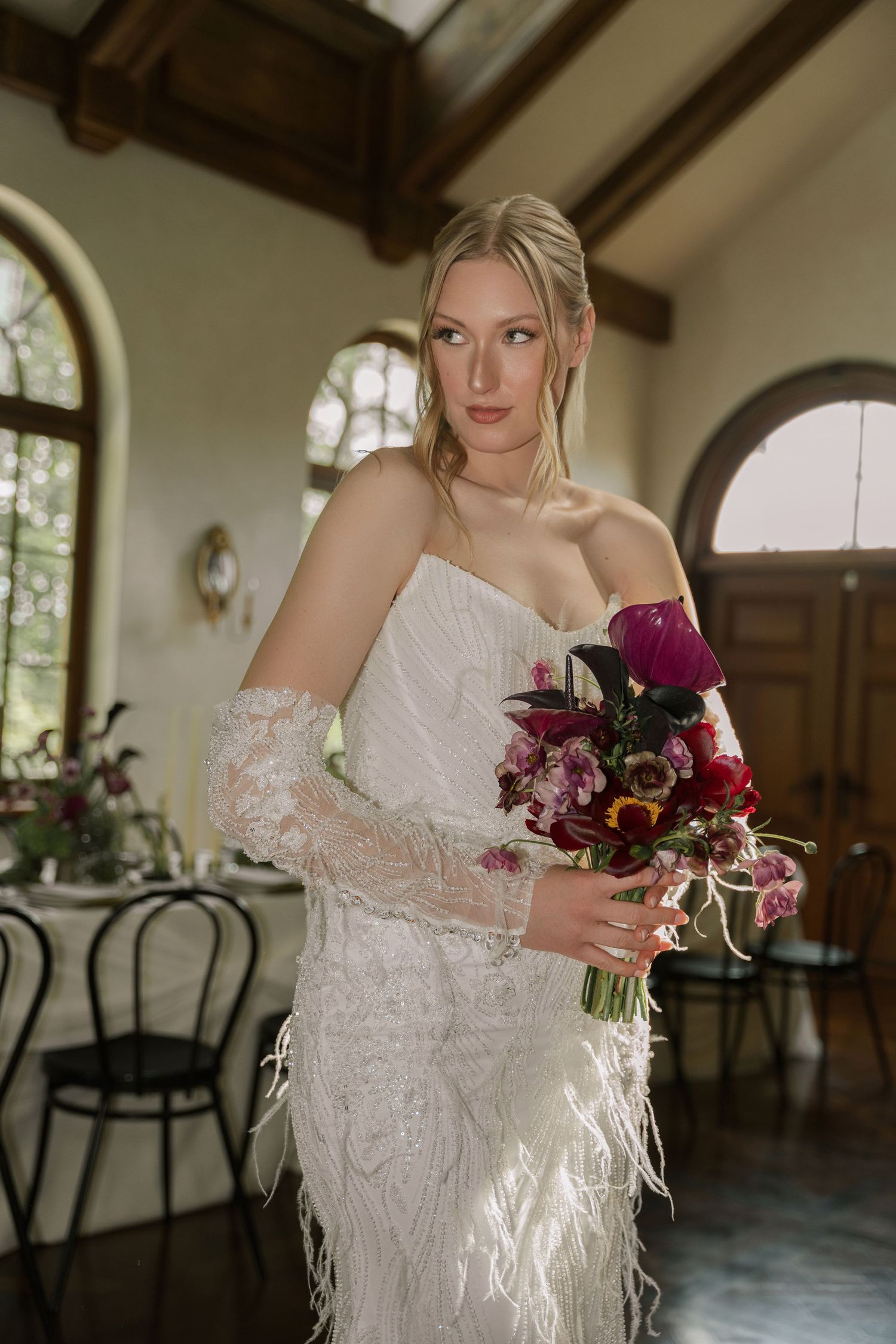 A bride in a feathered white gown holds a deep burgundy and pink bouquet in an elegant venue with wooden beams.