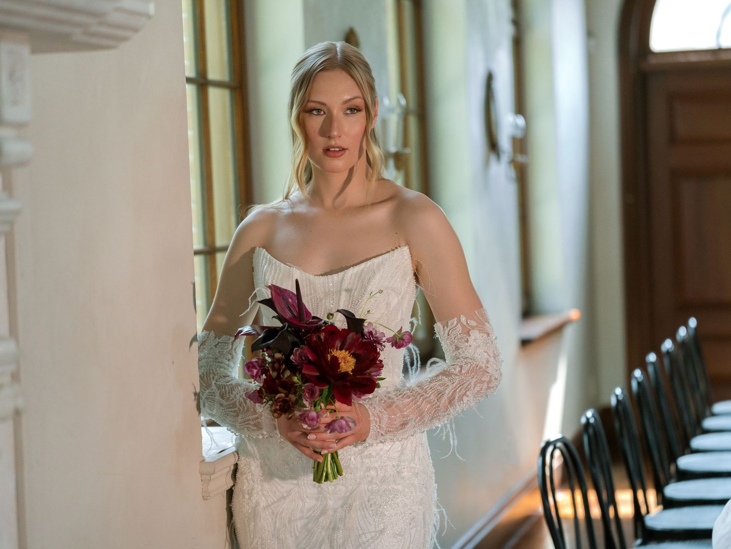 A bride in a lace off-shoulder wedding dress holds a deep burgundy and pink floral bouquet in an elegant hallway.