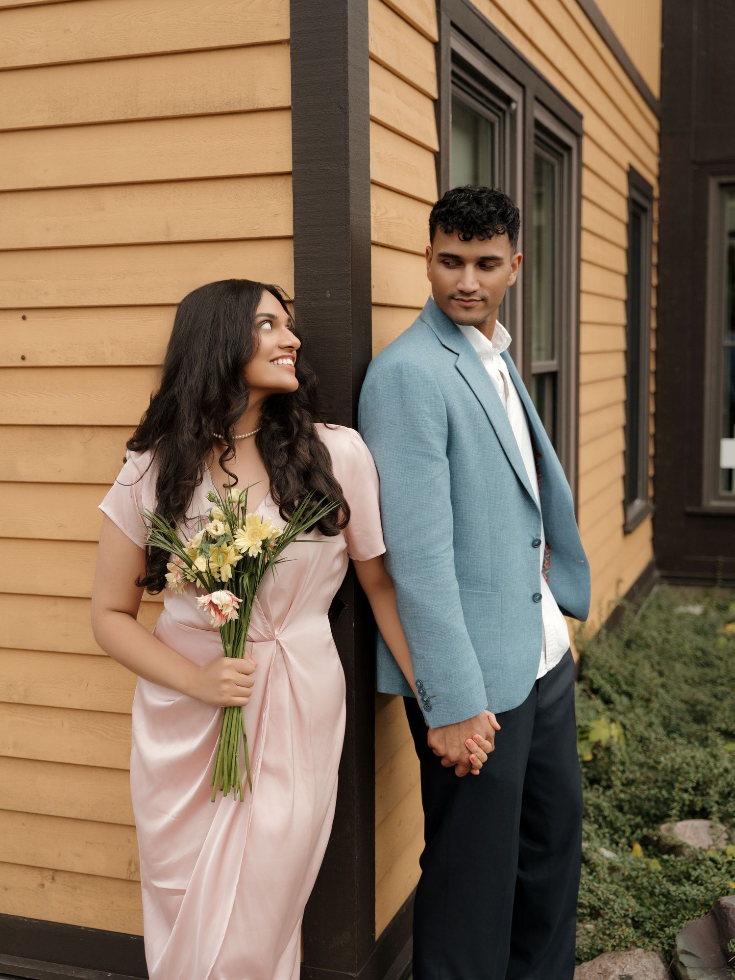 A couple in light pastel clothing holds hands while standing by a yellow building exterior with flowers.