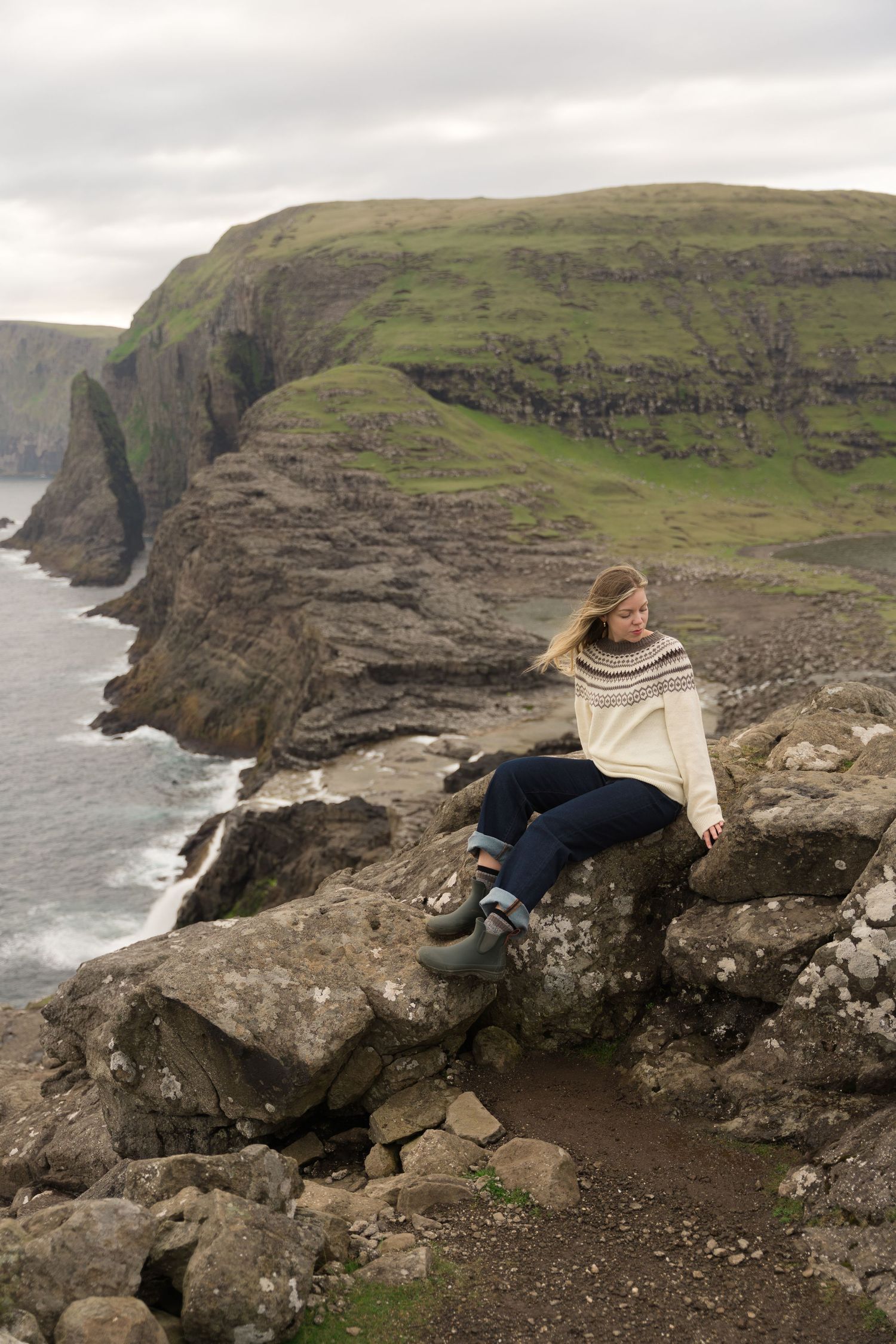 Woman sitting on coastal cliffs wearing a Nordic sweater, with dramatic green headlands and ocean views behind her.