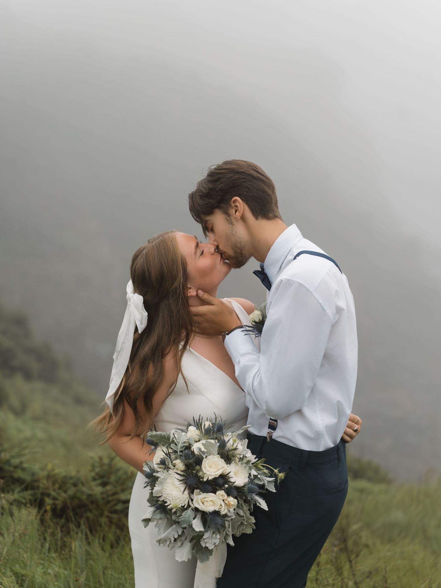 Newlyweds share a romantic kiss in a misty mountain setting with the bride holding a white and green floral bouquet.