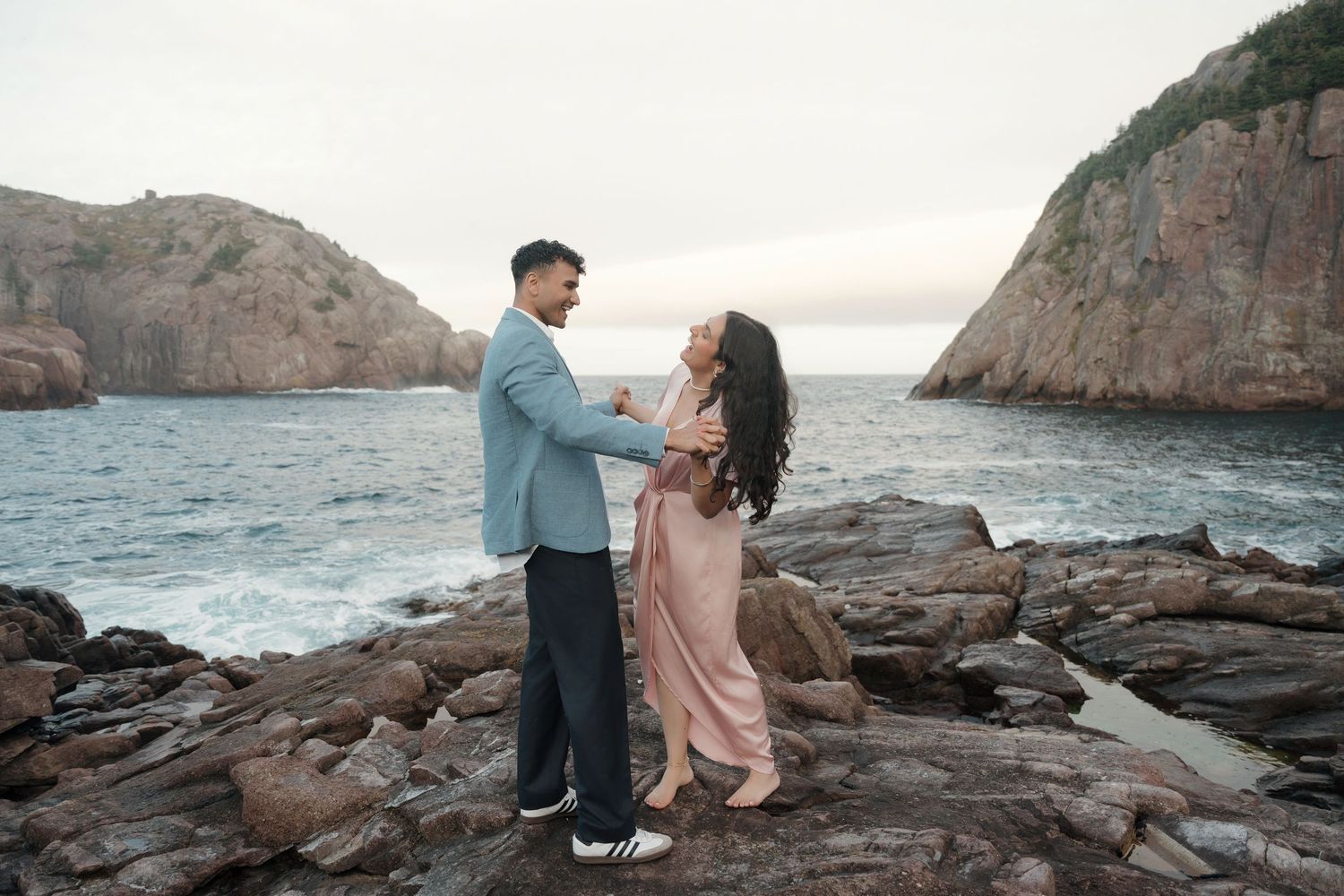 A couple shares tender moments along a rocky coastal landscape at sunset as waves crash against the shore.