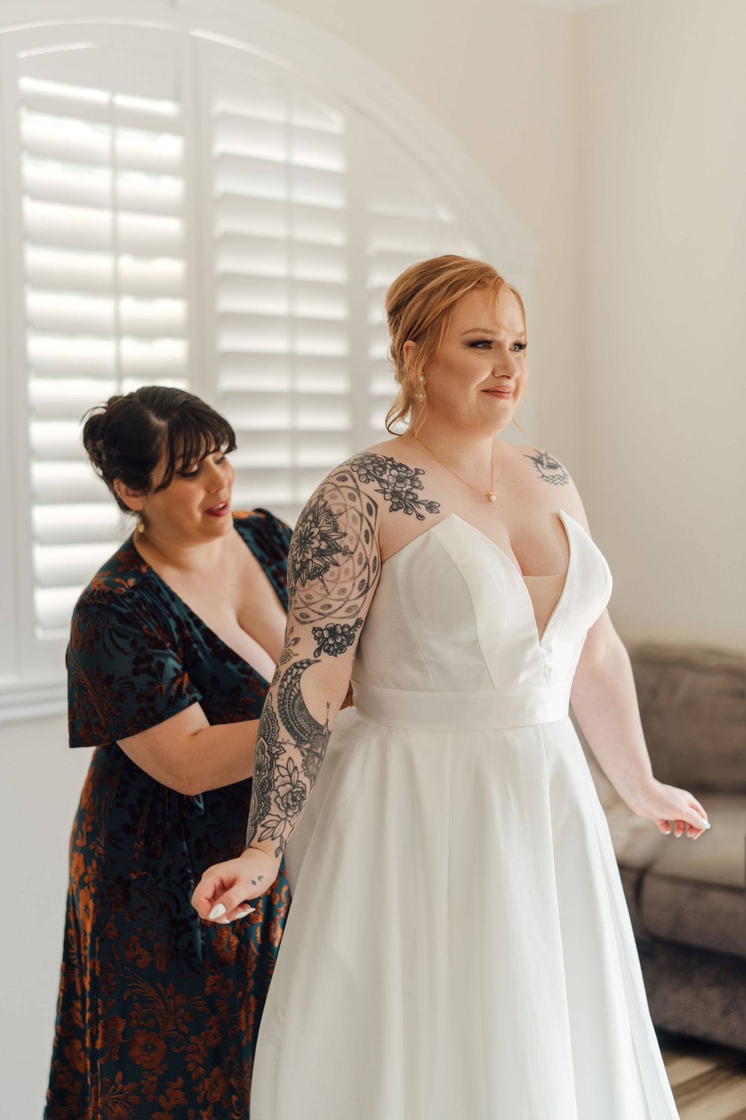 A bride in a white wedding dress stands while being helped with final adjustments.