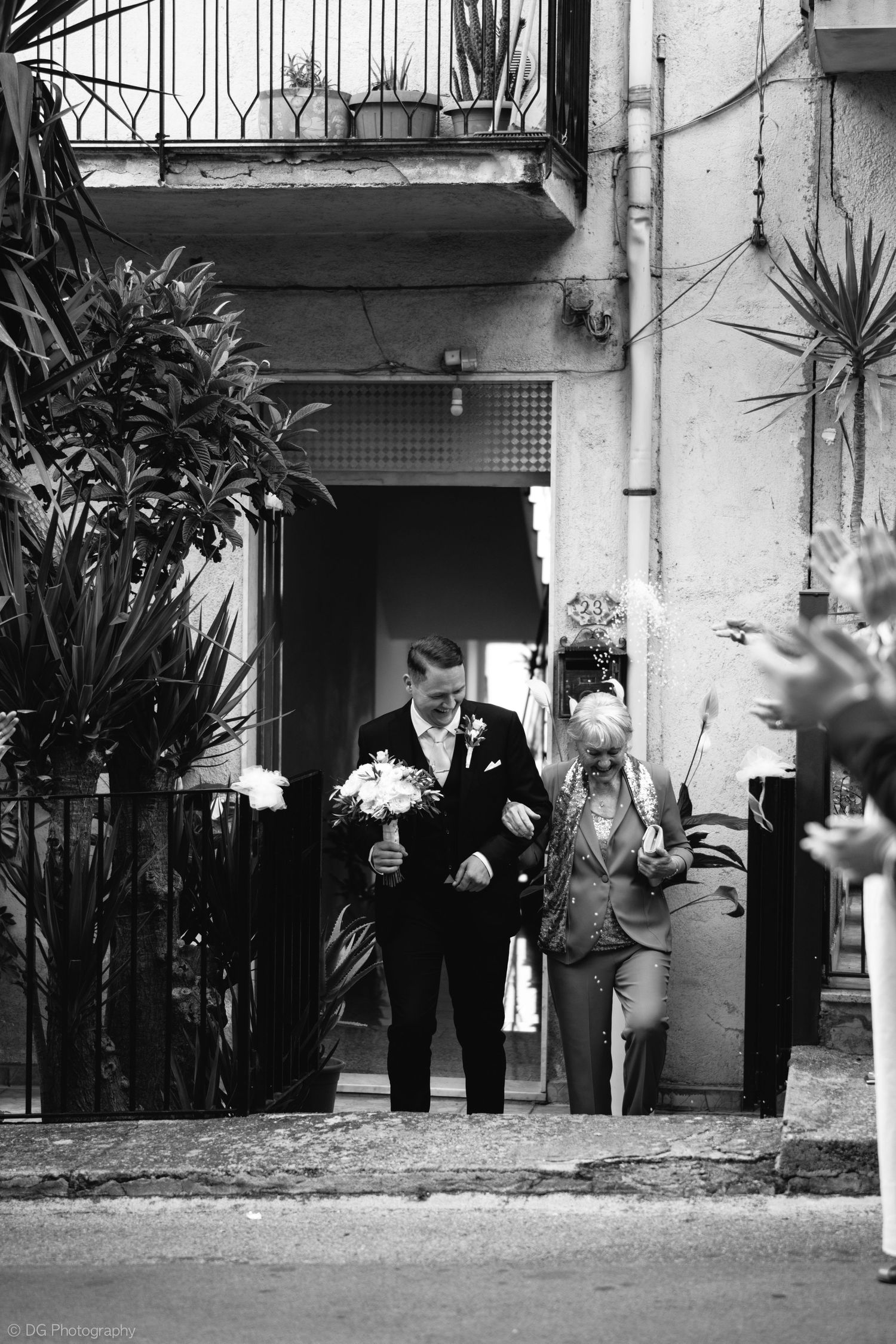 Black and white panoramic photo showing people in formal attire standing against a wall with plants and columns.