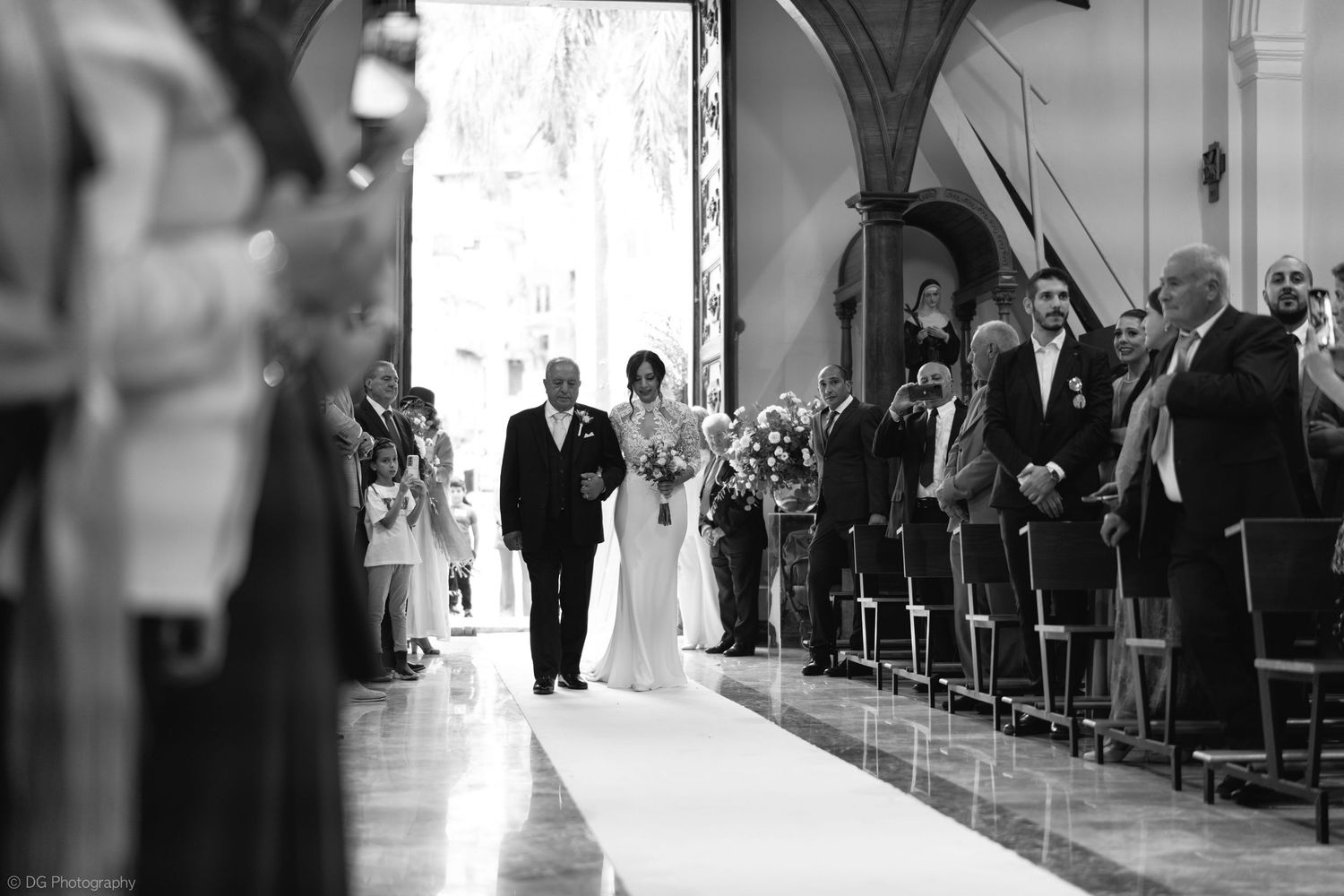 Wedding ceremony guests stand in church pews watching as bride and groom walk down the aisle together.
