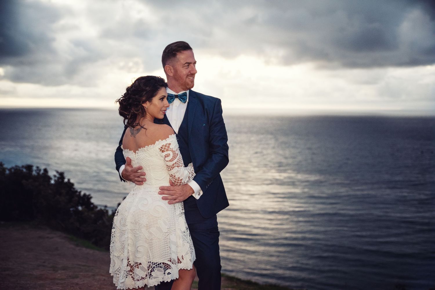 A romantic couple in wedding attire embrace at sunset overlooking a dramatic ocean view from a coastal cliff.
