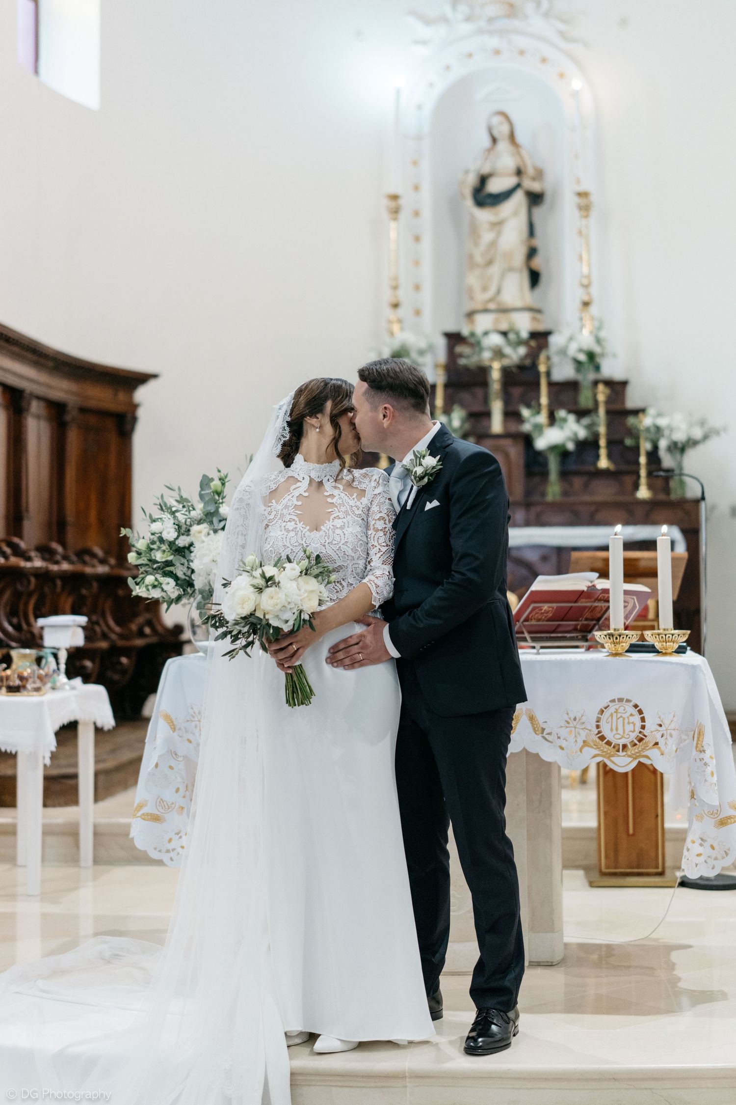 A couple shares their first dance in an elegant white church sanctuary decorated with floral arrangements and candles.