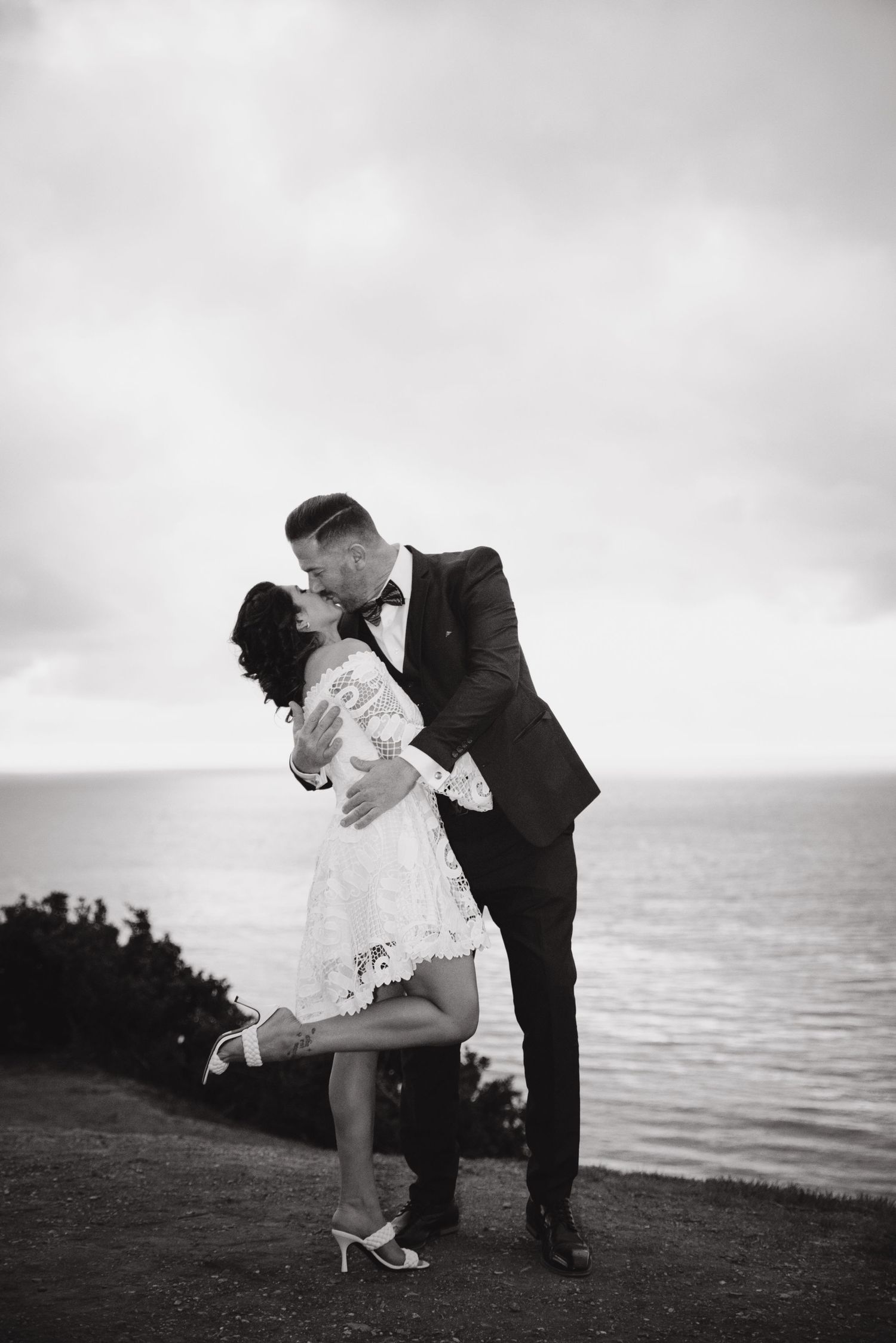 A romantic black and white photo of a couple embracing and kissing on a cliff overlooking the ocean.