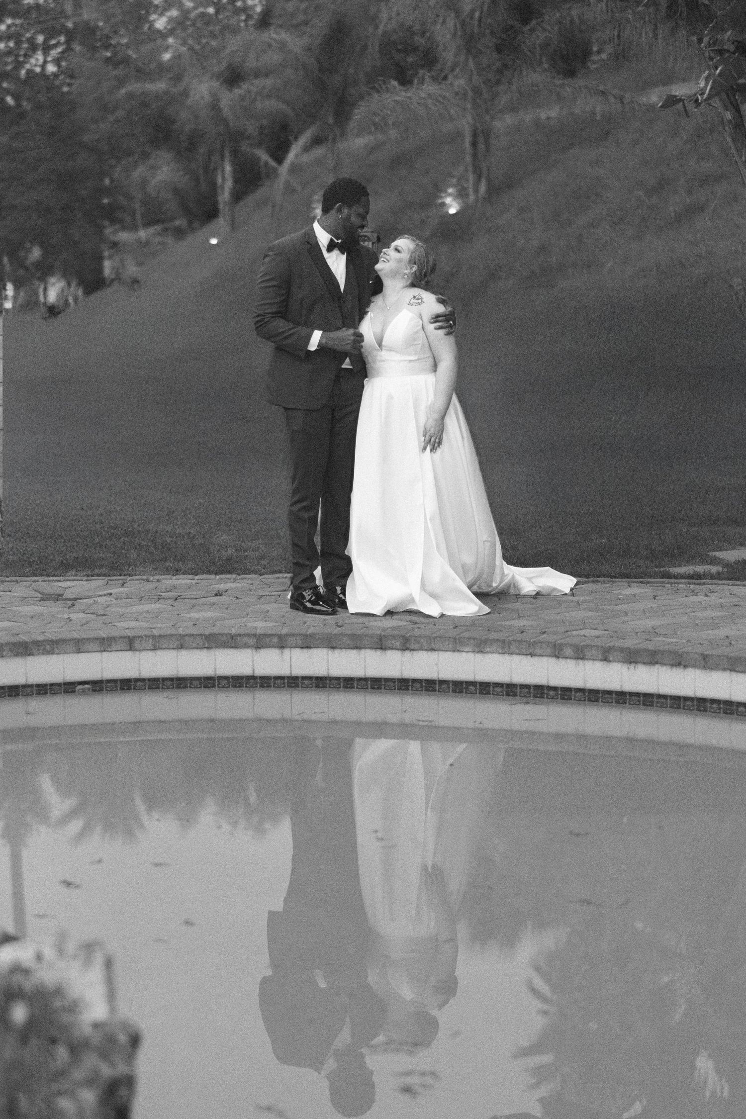 Newlyweds pose by a reflective pool in elegant black and white wedding attire.