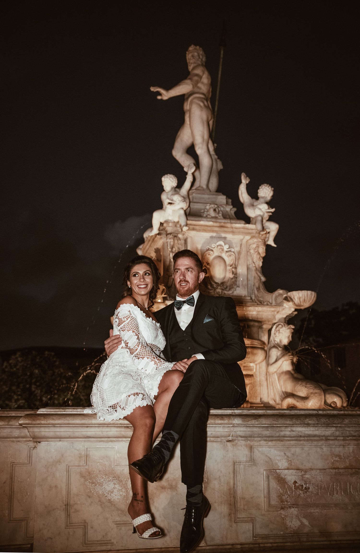 A romantic couple poses at night near an ornate stone fountain during their wedding celebration.