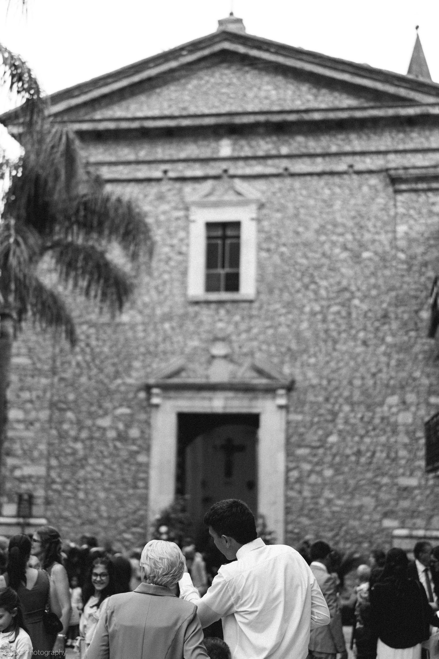 A crowd gathers outside a historic stone church building with traditional architecture and palm trees visible.
