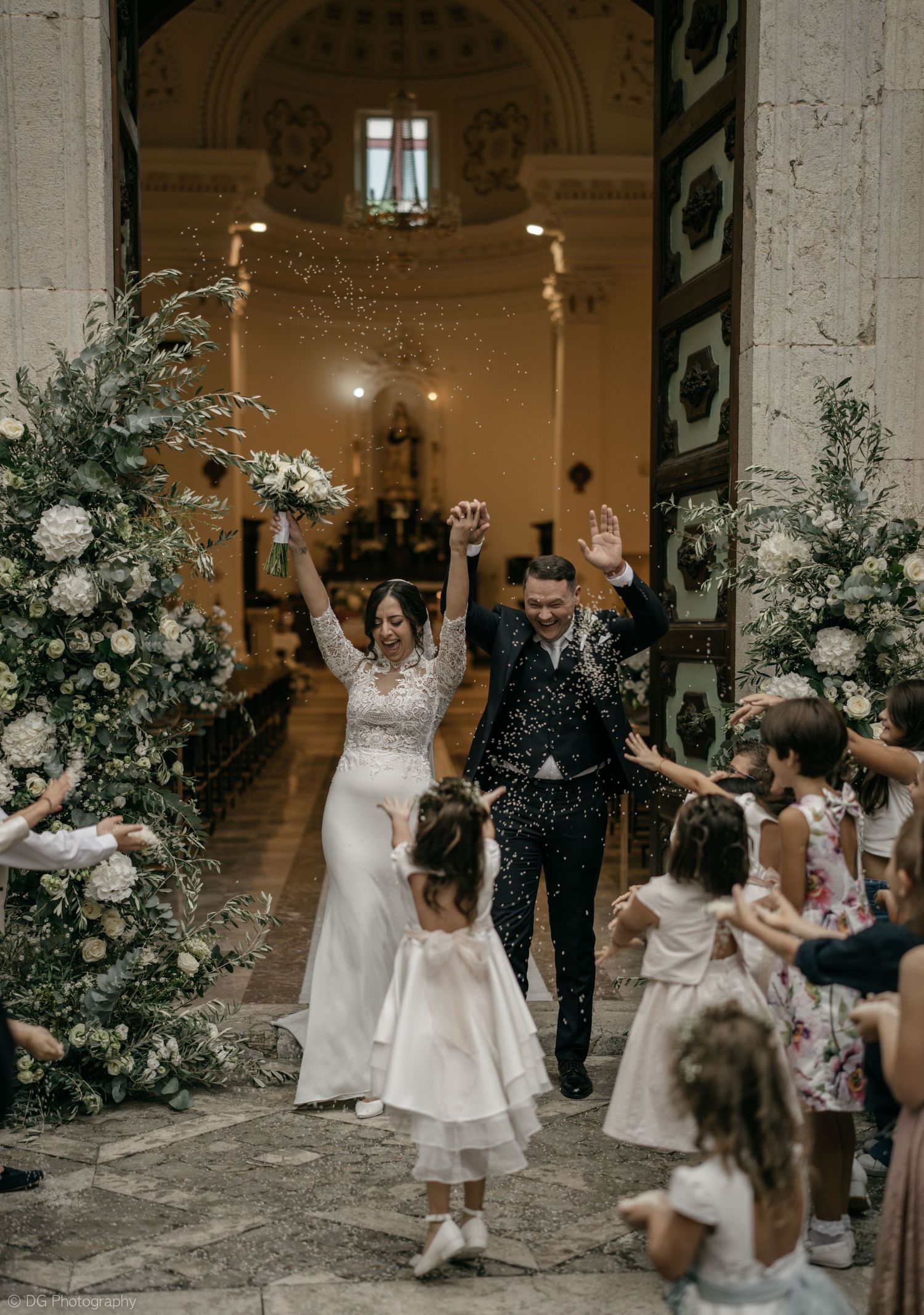 Newlyweds celebrate with joyful arms raised while exiting a church decorated with elegant white floral arrangements.