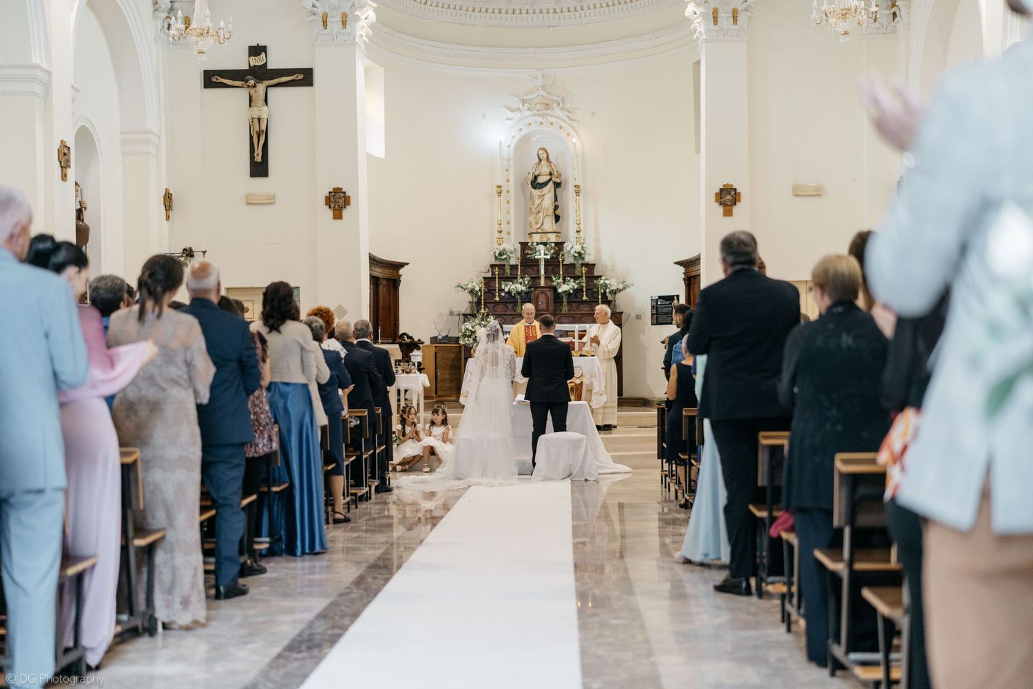 Wedding guests stand in elegant church pews during a traditional Catholic ceremony in a bright white sanctuary.