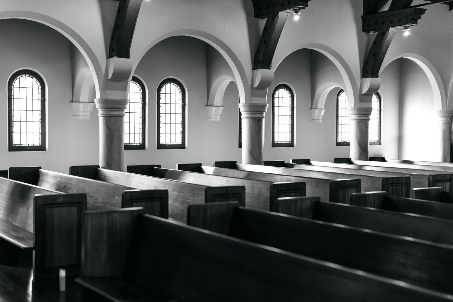 Black and white interior photograph of an empty church sanctuary with rows of wooden pews and arched windows.