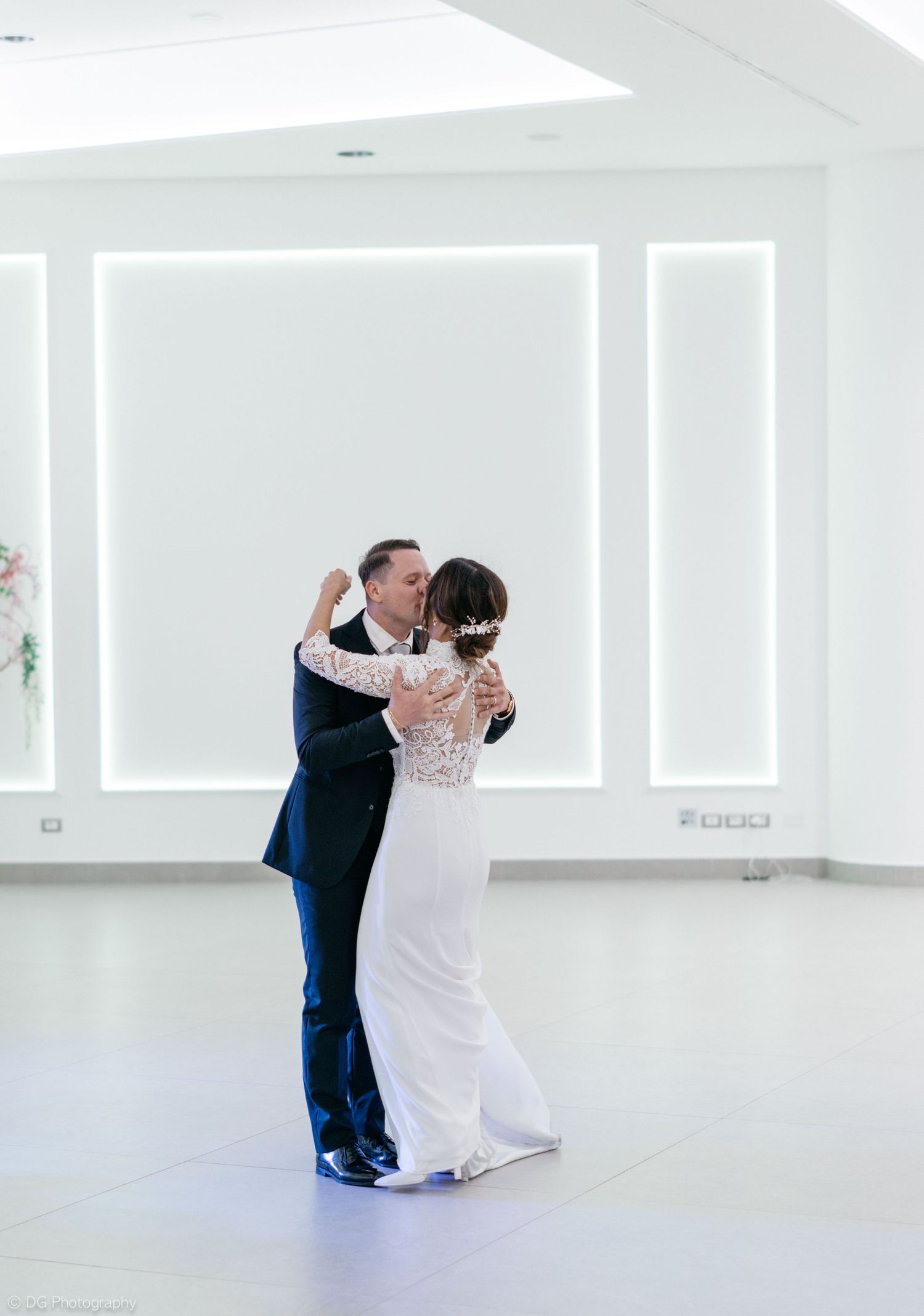 A couple shares a romantic dance in a bright white modern space with floor-to-ceiling windows.