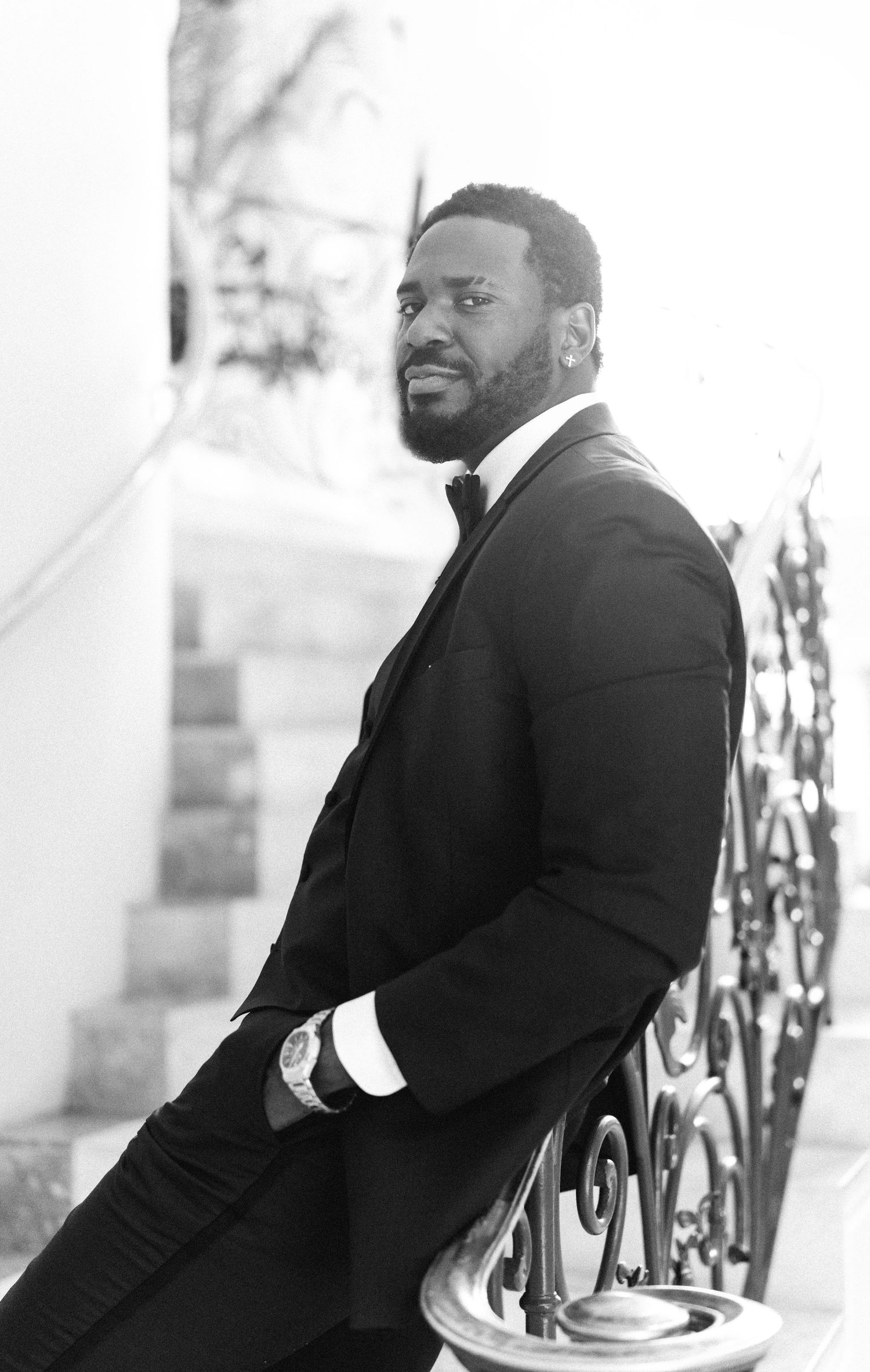 A groom in a black tuxedo poses against an elegant white wall with iron railings.